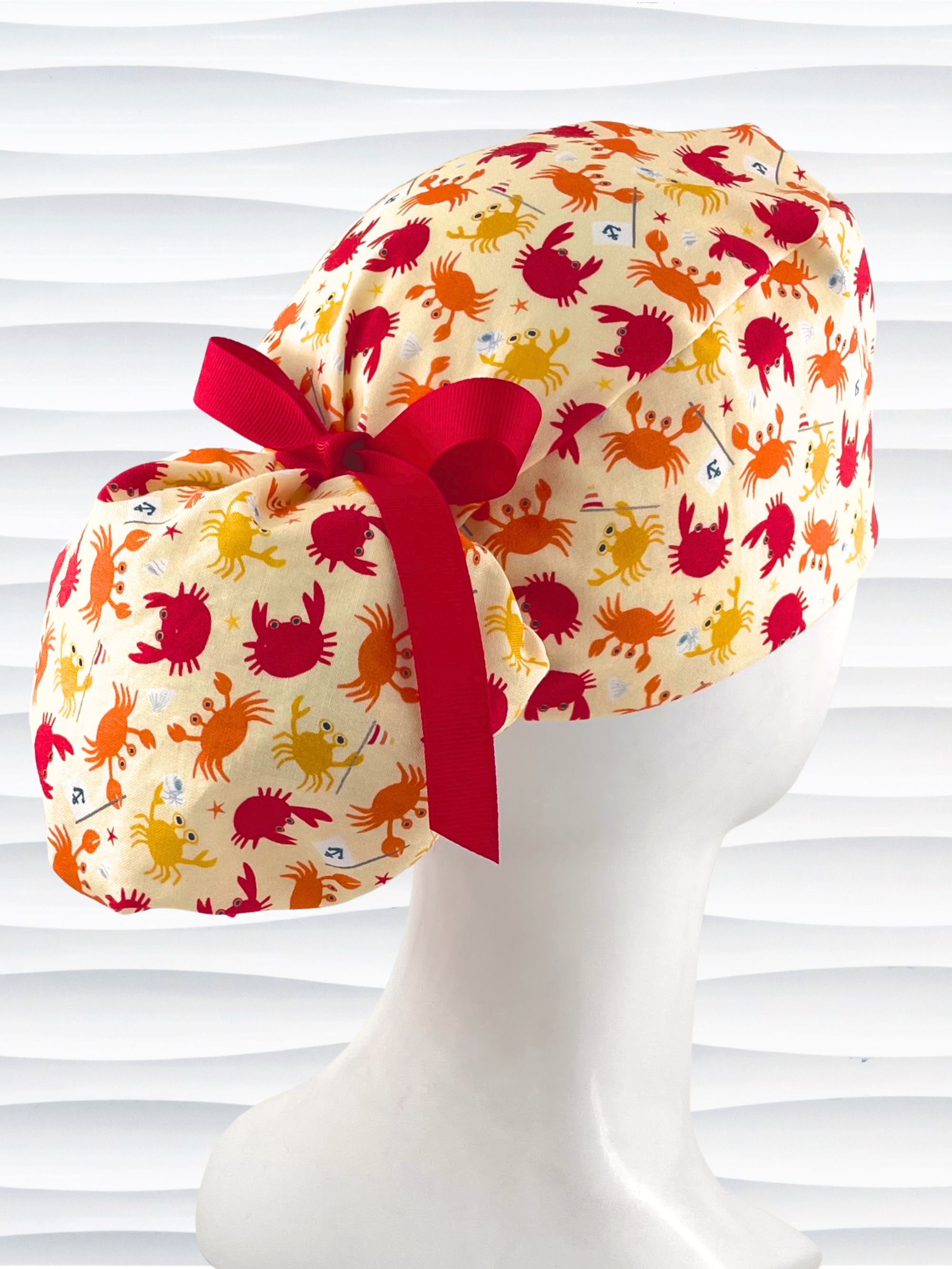 Colorful crab print surgical scrub cap with red ribbon tie, featuring orange, red, and yellow cartoon crabs on cream fabric, displayed on mannequin head – handmade nurse and doctor scrub hat by JensCaps