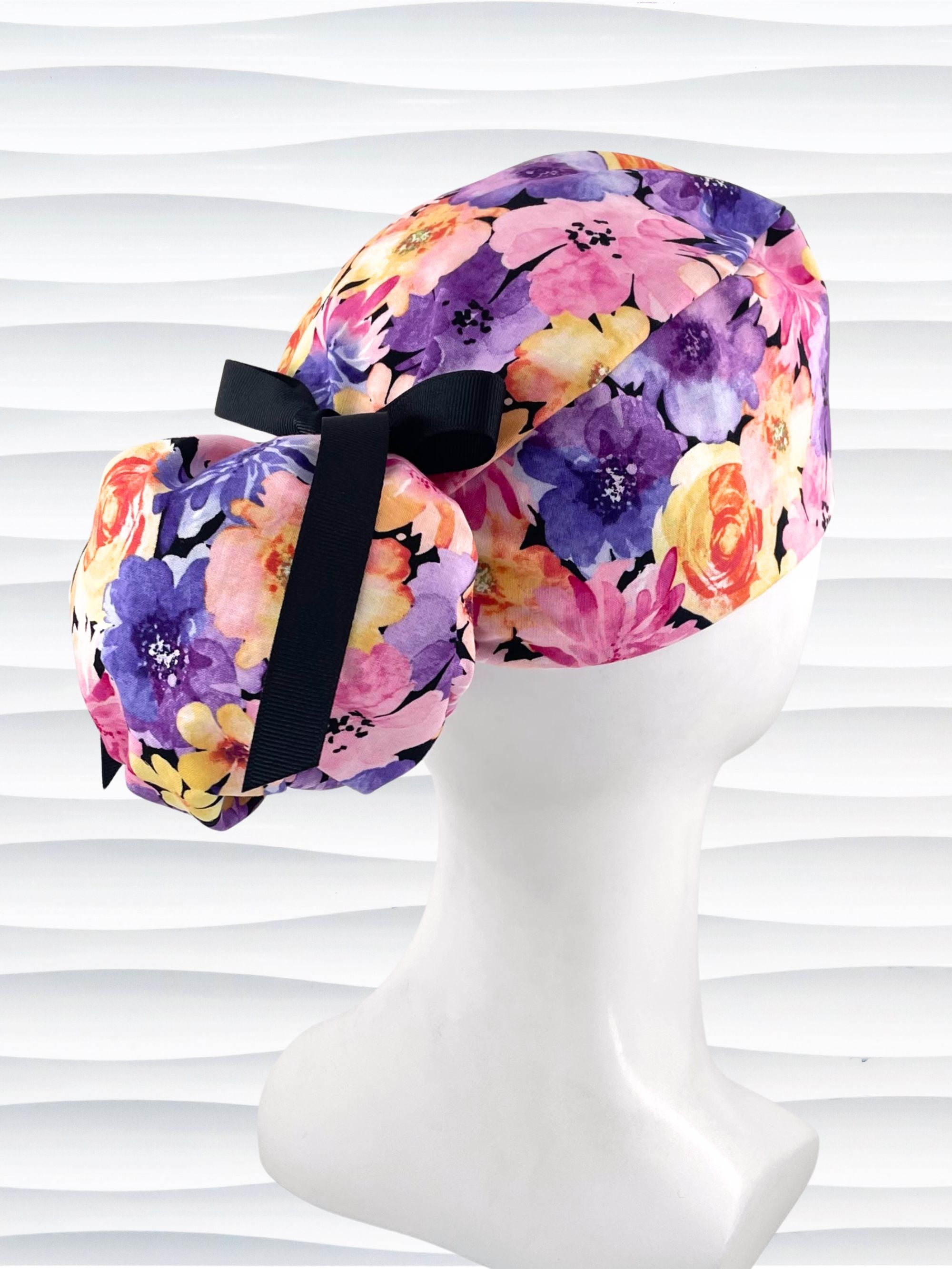Yellow, pink, and purple floral-patterned Ponytail scrub cap by JensCaps with a black grosgrain ribbon on a mannequin head against a white background
