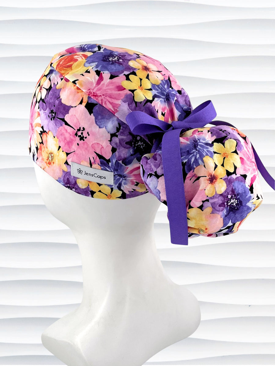 Yellow, pink, and purple floral-patterned Ponytail scrub cap by JensCaps with a purple grosgrain ribbon on a mannequin head against a white background