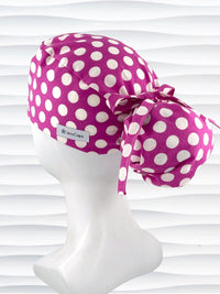 Ponytail style surgical scrub cap by JensCaps with large white retro inspired polka dots on a rasberry colored cotton fabric, and finished with a ribbon tie.