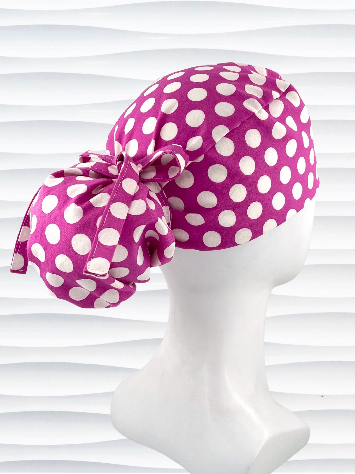 Ponytail style surgical scrub cap by JensCaps with large white retro inspired polka dots on a rasberry colored cotton fabric, and finished with a ribbon tie.