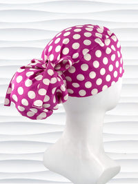 Ponytail style surgical scrub cap by JensCaps with large white retro inspired polka dots on a rasberry colored cotton fabric, and finished with a ribbon tie.