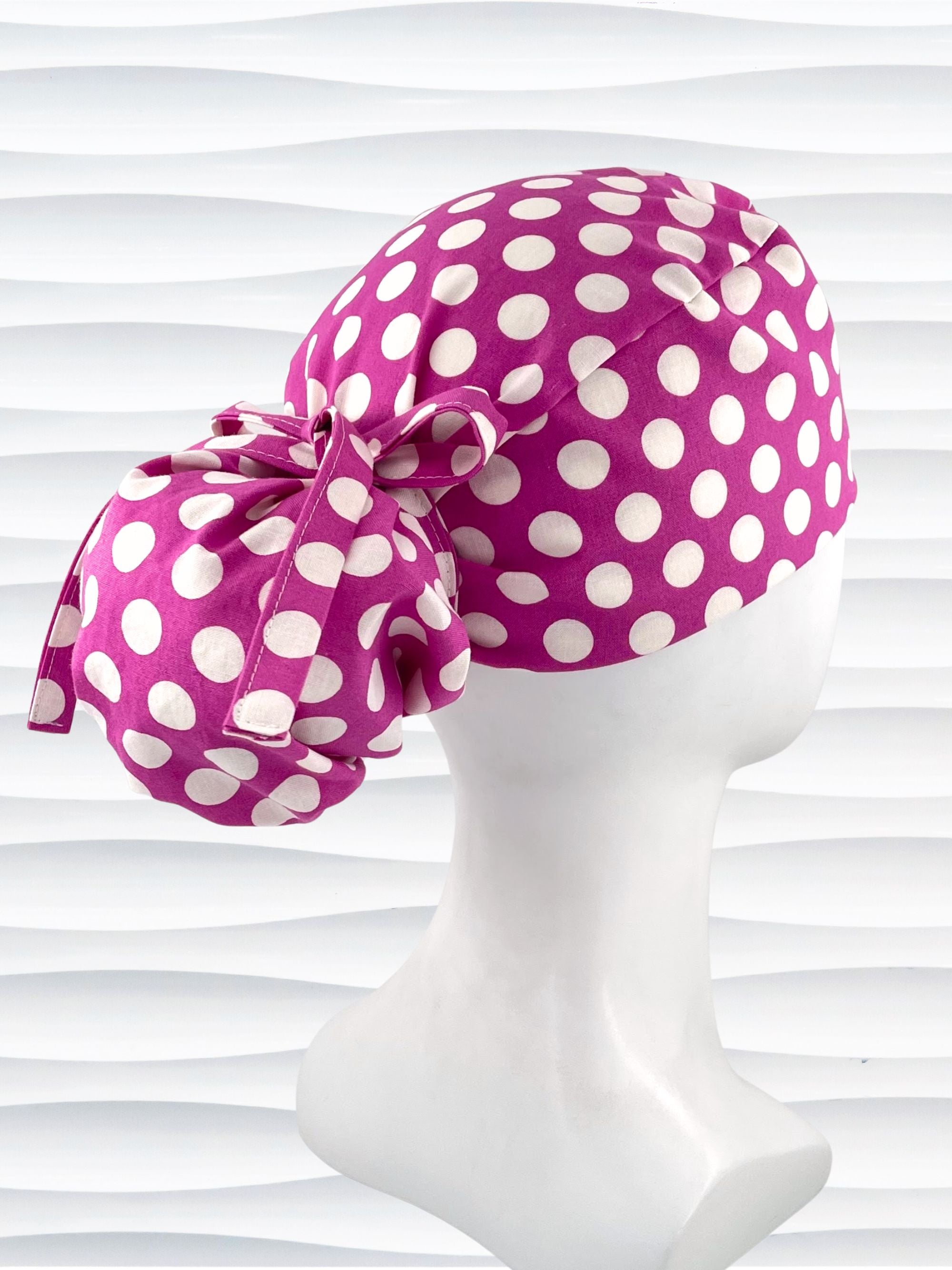 Ponytail style surgical scrub cap by JensCaps with large white retro inspired polka dots on a rasberry colored cotton fabric, and finished with a ribbon tie.