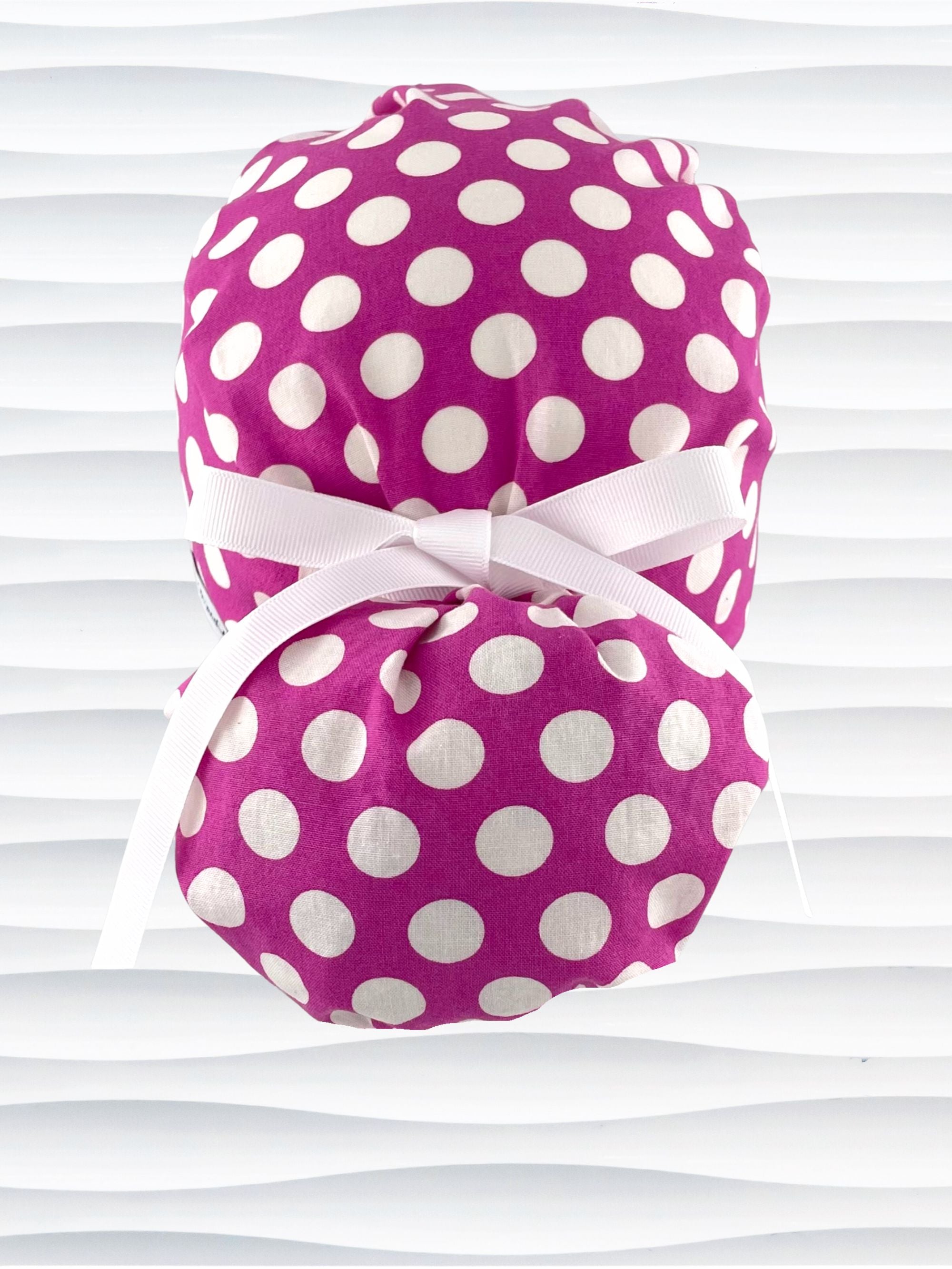 Ponytail style surgical scrub cap by JensCaps with large white retro inspired polka dots on a rasberry colored cotton fabric, and finished with a ribbon tie.