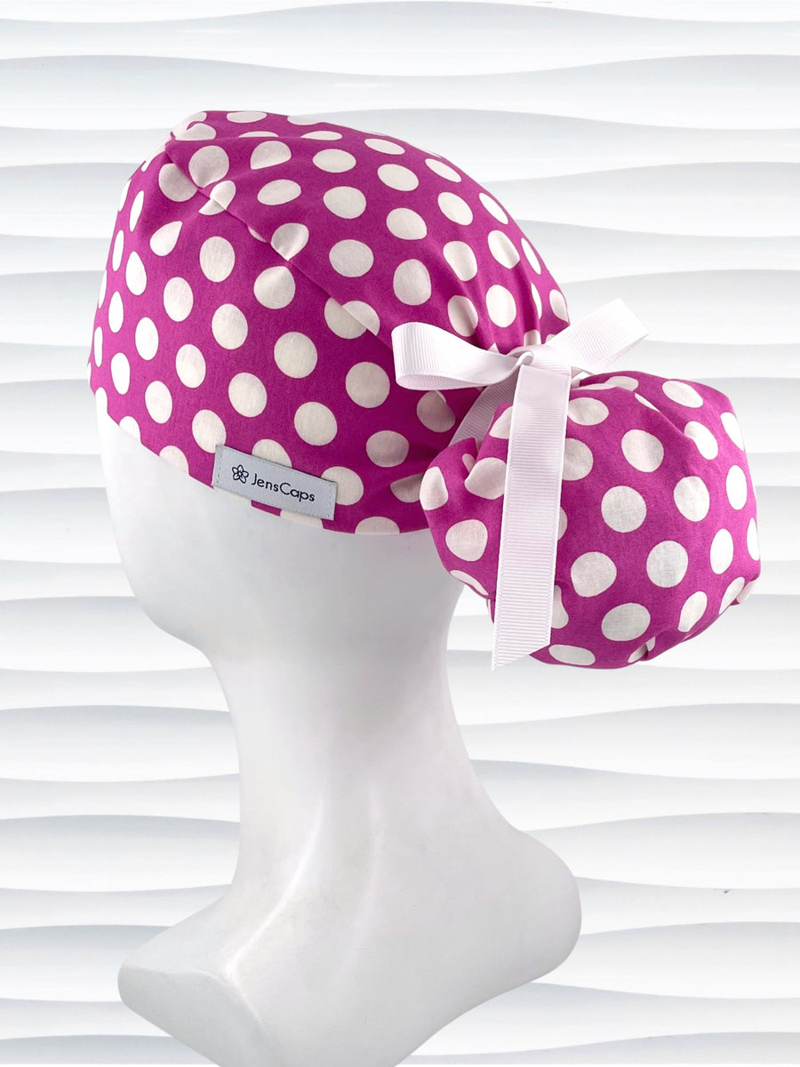 Ponytail style surgical scrub cap by JensCaps with large white retro inspired polka dots on a rasberry colored cotton fabric, and finished with a ribbon tie.