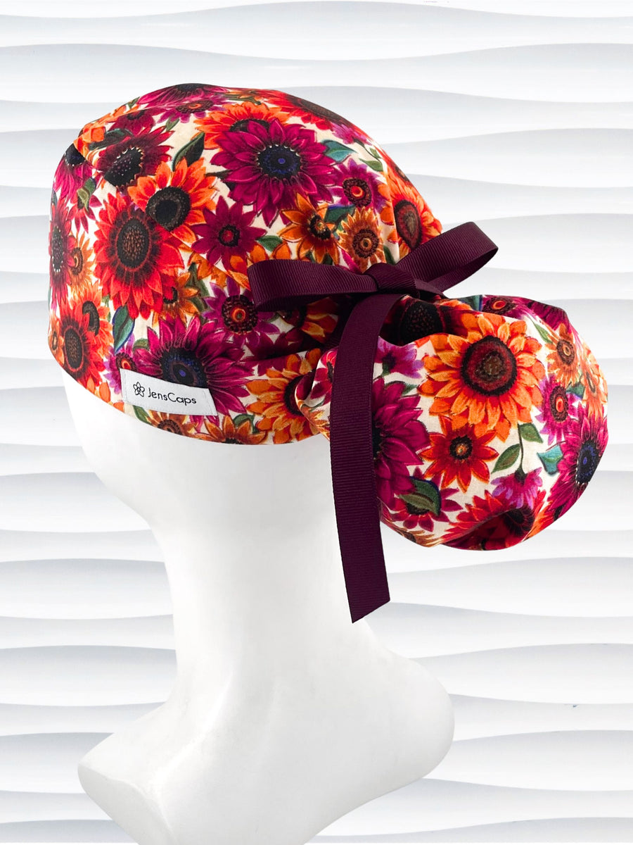 Ponytail style surgical scrub cap with sunflowers in rich reds, pinks, and orange all over a white background and finished with ribbon ties.