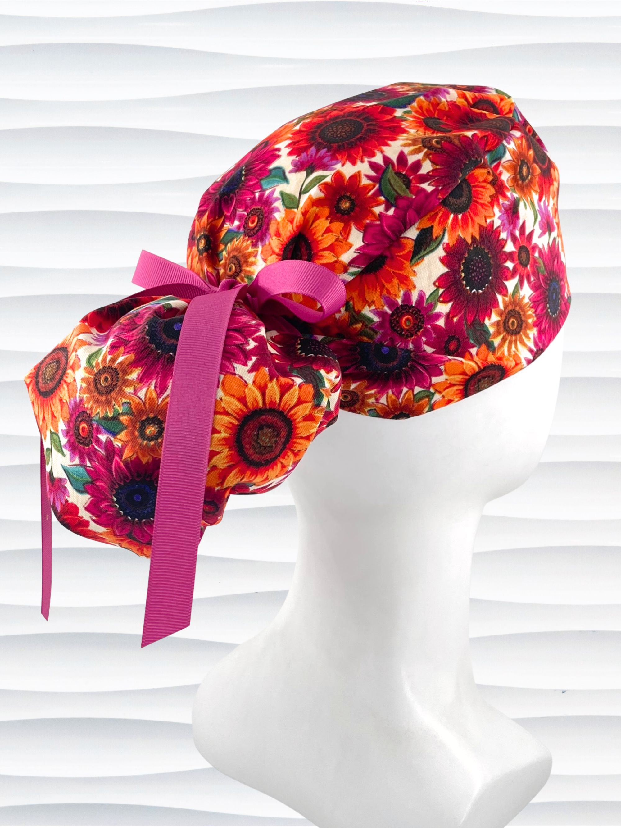 Ponytail style surgical scrub cap with sunflowers in rich reds, pinks, and orange all over a white background and finished with ribbon ties.
