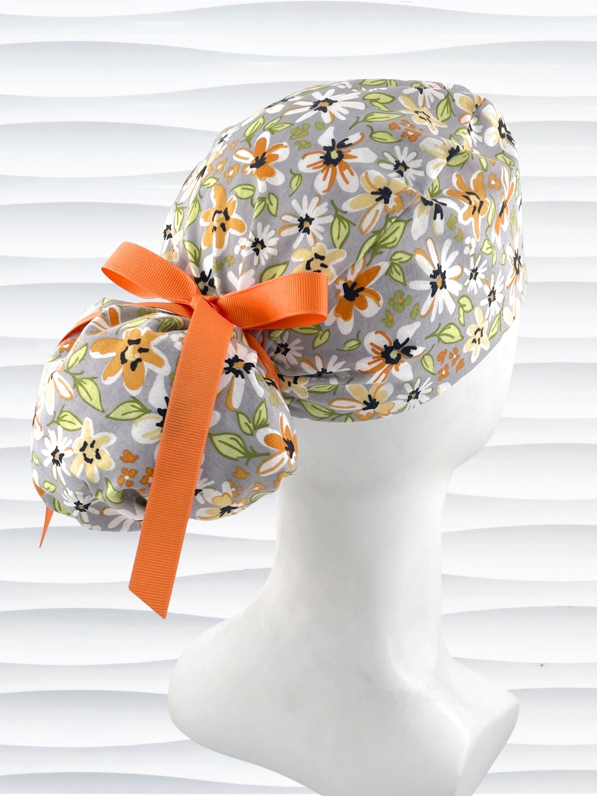 Ponytail style surgical scrub cap by JensCaps with yellow, white, and black flowers on a gray cotton fabric, finished with a grosgrain ribbon, and displayed on a mannequin head.