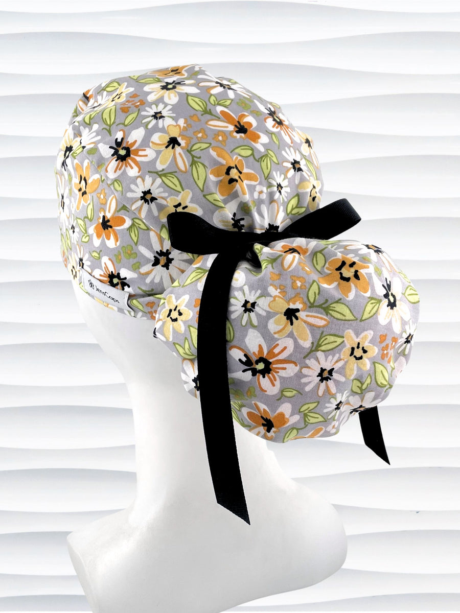 Ponytail style surgical scrub cap by JensCaps with yellow, white, and black flowers on a gray cotton fabric, finished with a grosgrain ribbon, and displayed on a mannequin head.
