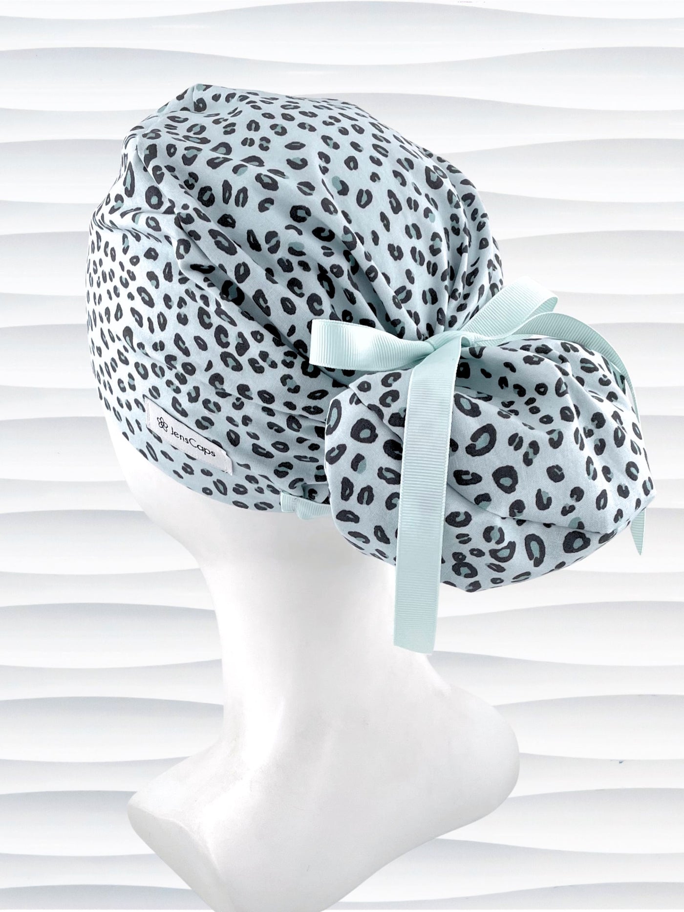 Euro Pony style surgical scrub cap by JensCaps with a leopard print on light teal cotton fabric, tied with a ribbon, and displayed on a mannequin head.