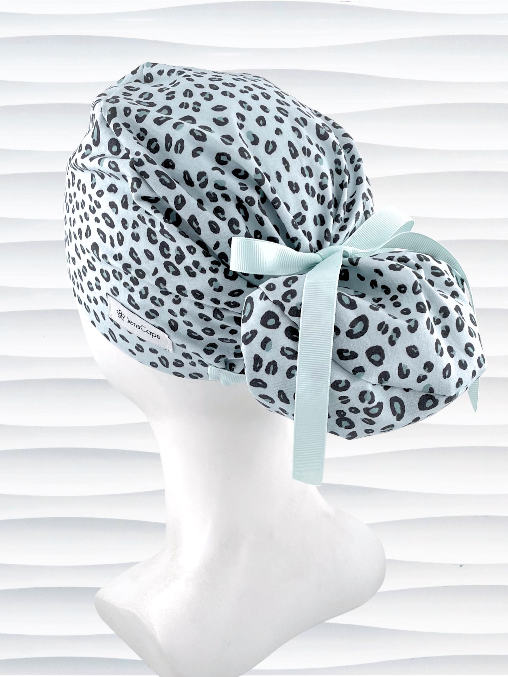 Euro Pony style surgical scrub cap by JensCaps with a leopard print on light teal cotton fabric, tied with a ribbon, and displayed on a mannequin head.