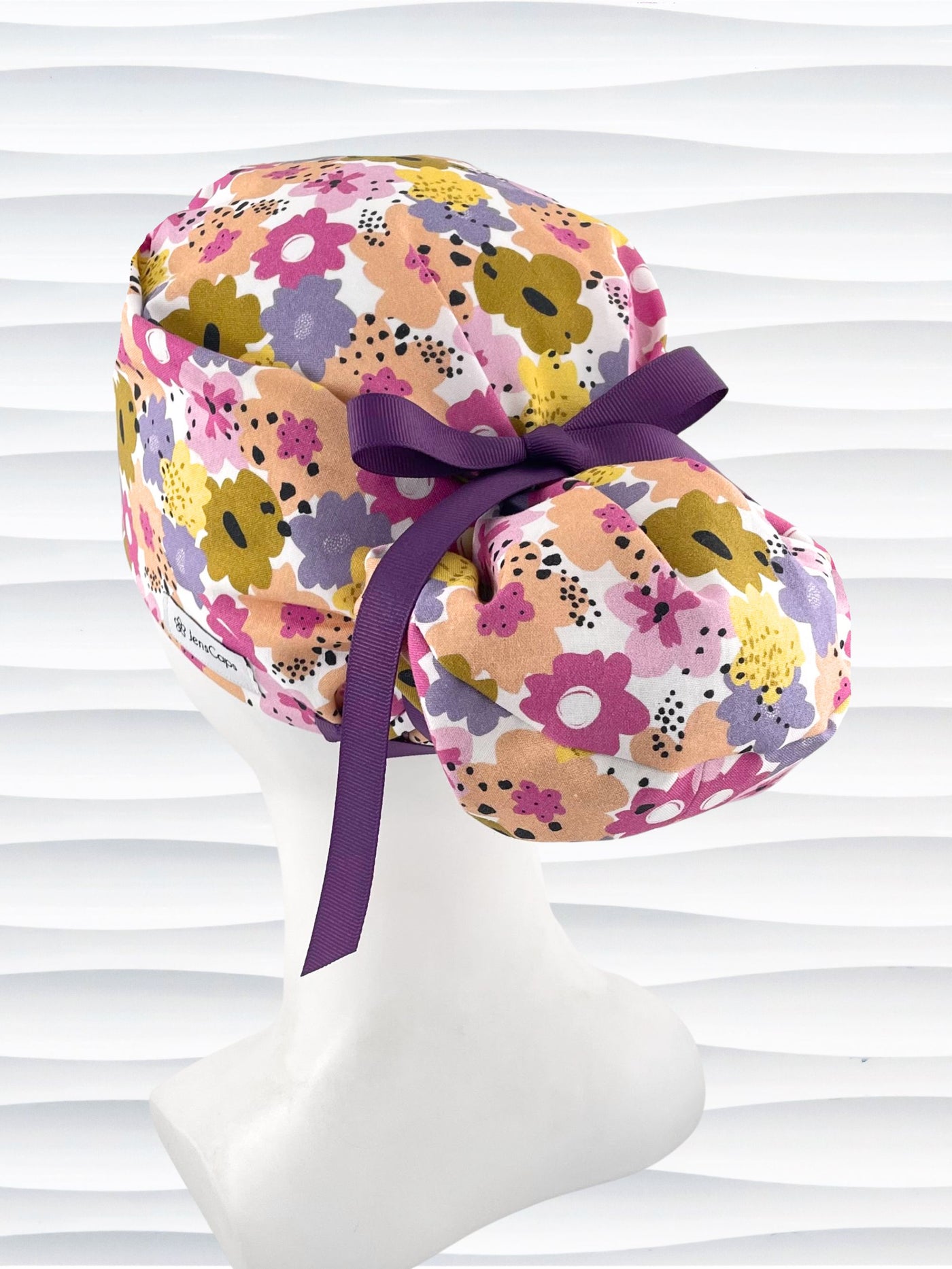 Euro Pony style surgical scrub cap by JensCaps with pink, purple, yellow, and green flowers all over white cotton fabric, tied with a ribbon and displayed on a mannequin head.
