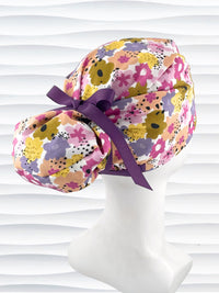 Euro Pony style surgical scrub cap by JensCaps with pink, purple, yellow, and green flowers all over white cotton fabric, tied with a ribbon and displayed on a mannequin head.