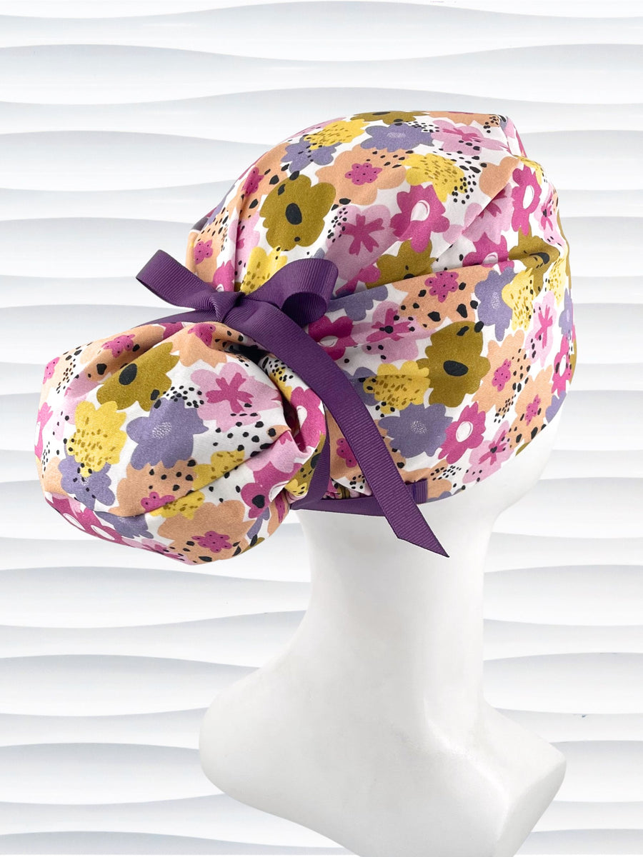 Euro Pony style surgical scrub cap by JensCaps with pink, purple, yellow, and green flowers all over white cotton fabric, tied with a ribbon and displayed on a mannequin head.