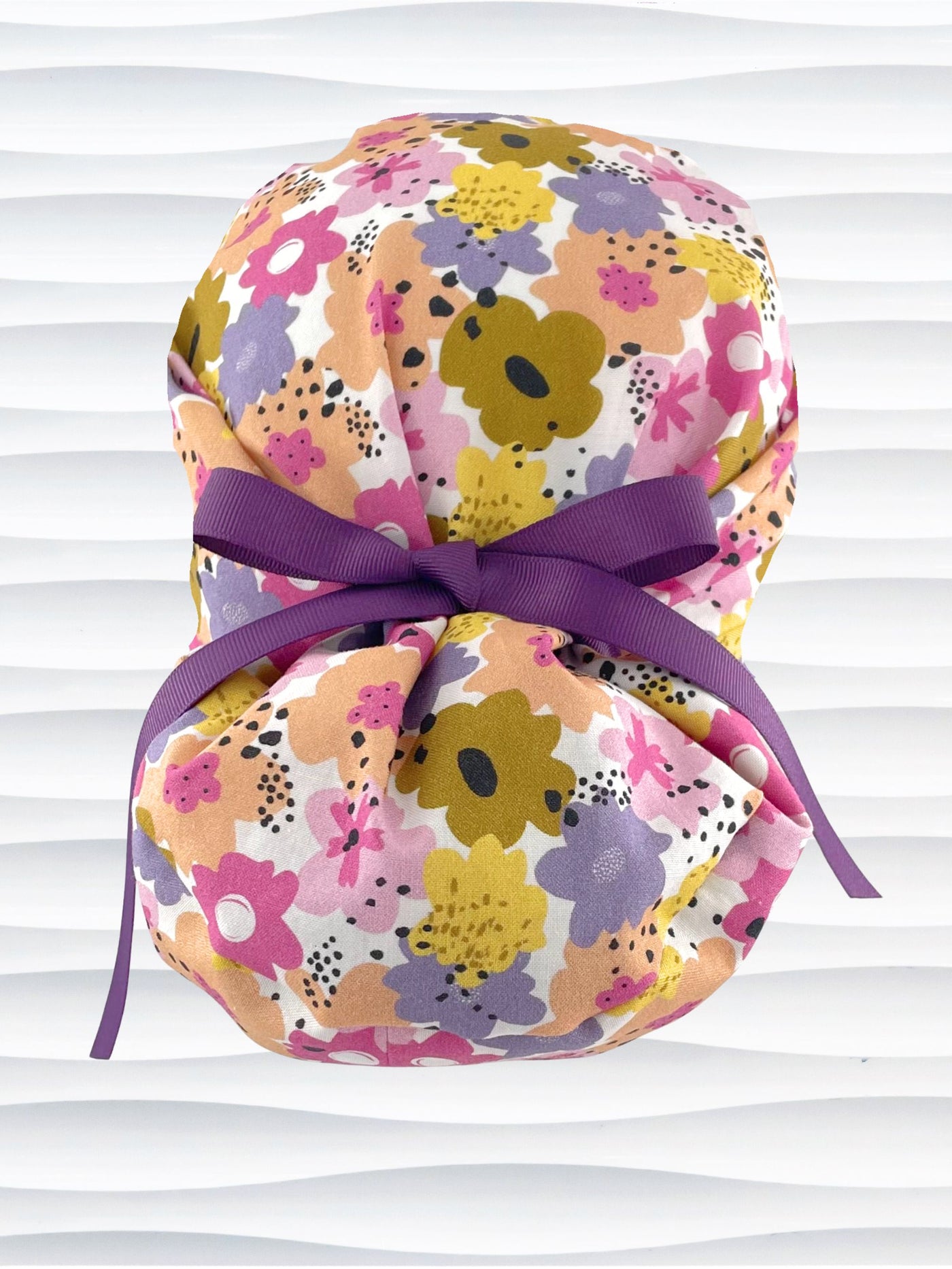 Euro Pony style surgical scrub cap by JensCaps with pink, purple, yellow, and green flowers all over white cotton fabric, tied with a ribbon and displayed on a mannequin head.