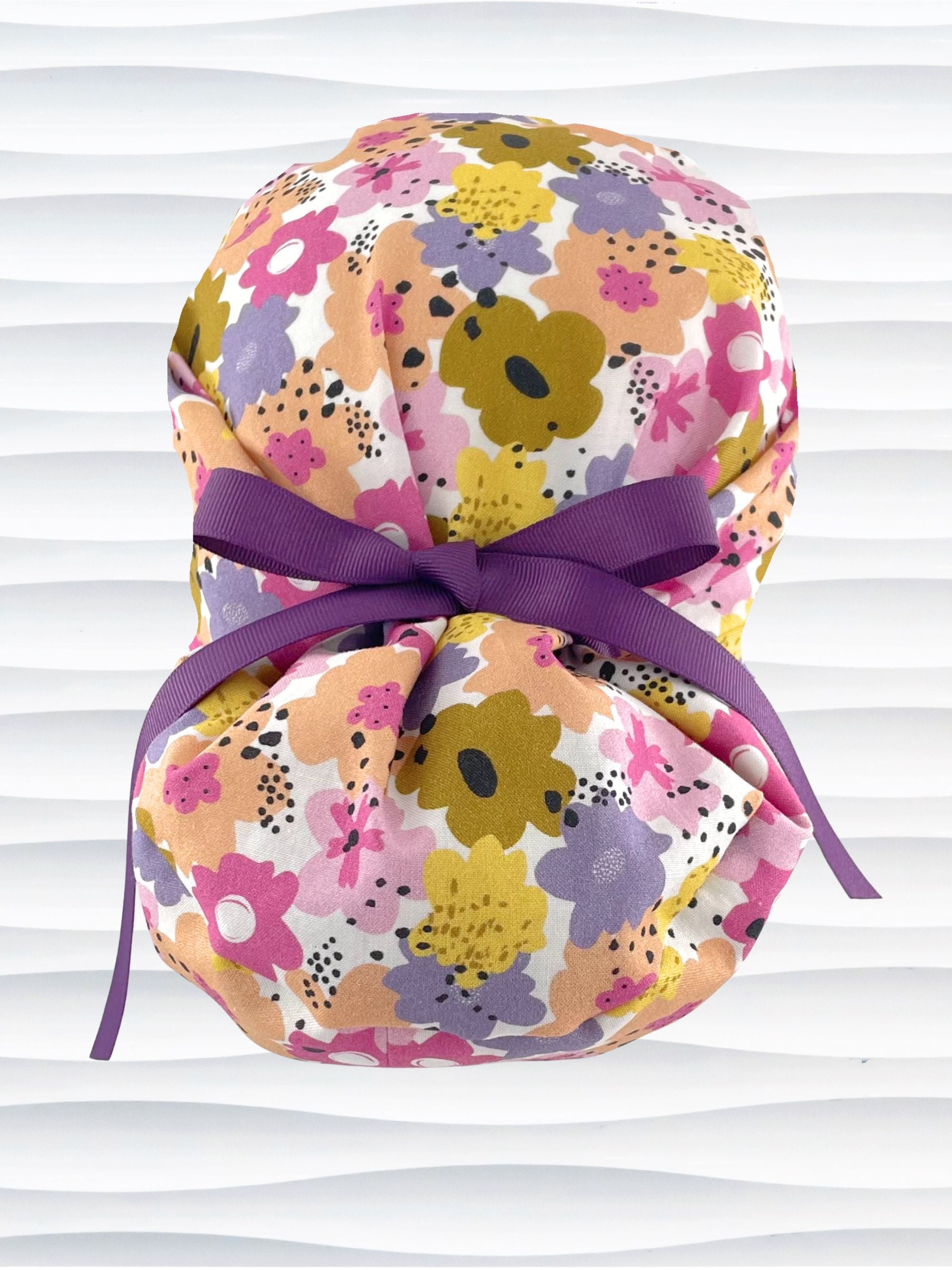 Euro Pony style surgical scrub cap by JensCaps with pink, purple, yellow, and green flowers all over white cotton fabric, tied with a ribbon and displayed on a mannequin head.