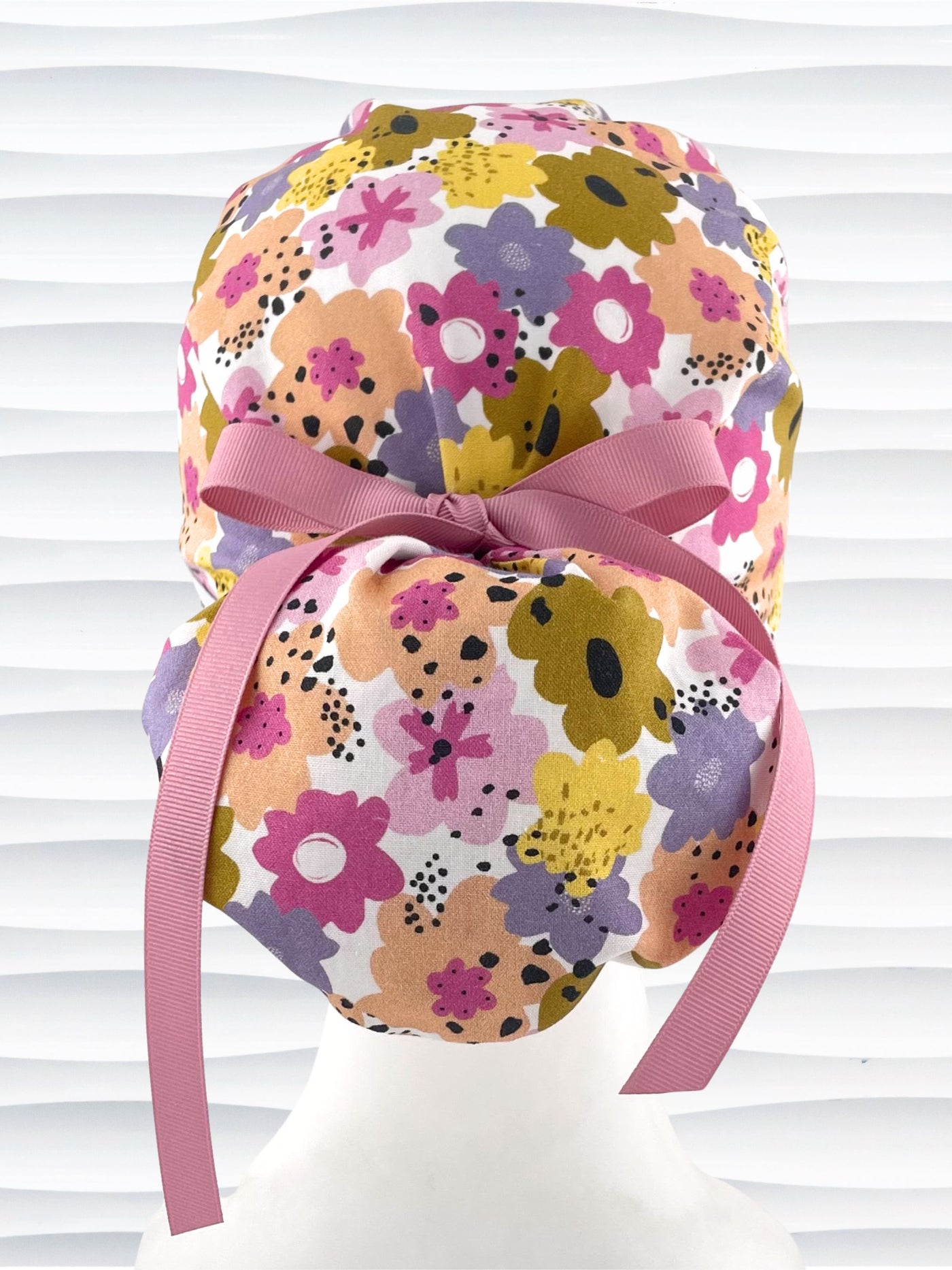 Euro Pony style surgical scrub cap by JensCaps with pink, purple, yellow, and green flowers all over white cotton fabric, tied with a ribbon and displayed on a mannequin head.
