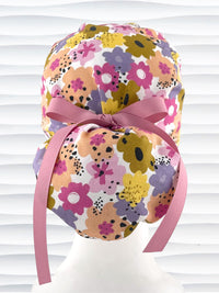 Euro Pony style surgical scrub cap by JensCaps with pink, purple, yellow, and green flowers all over white cotton fabric, tied with a ribbon and displayed on a mannequin head.