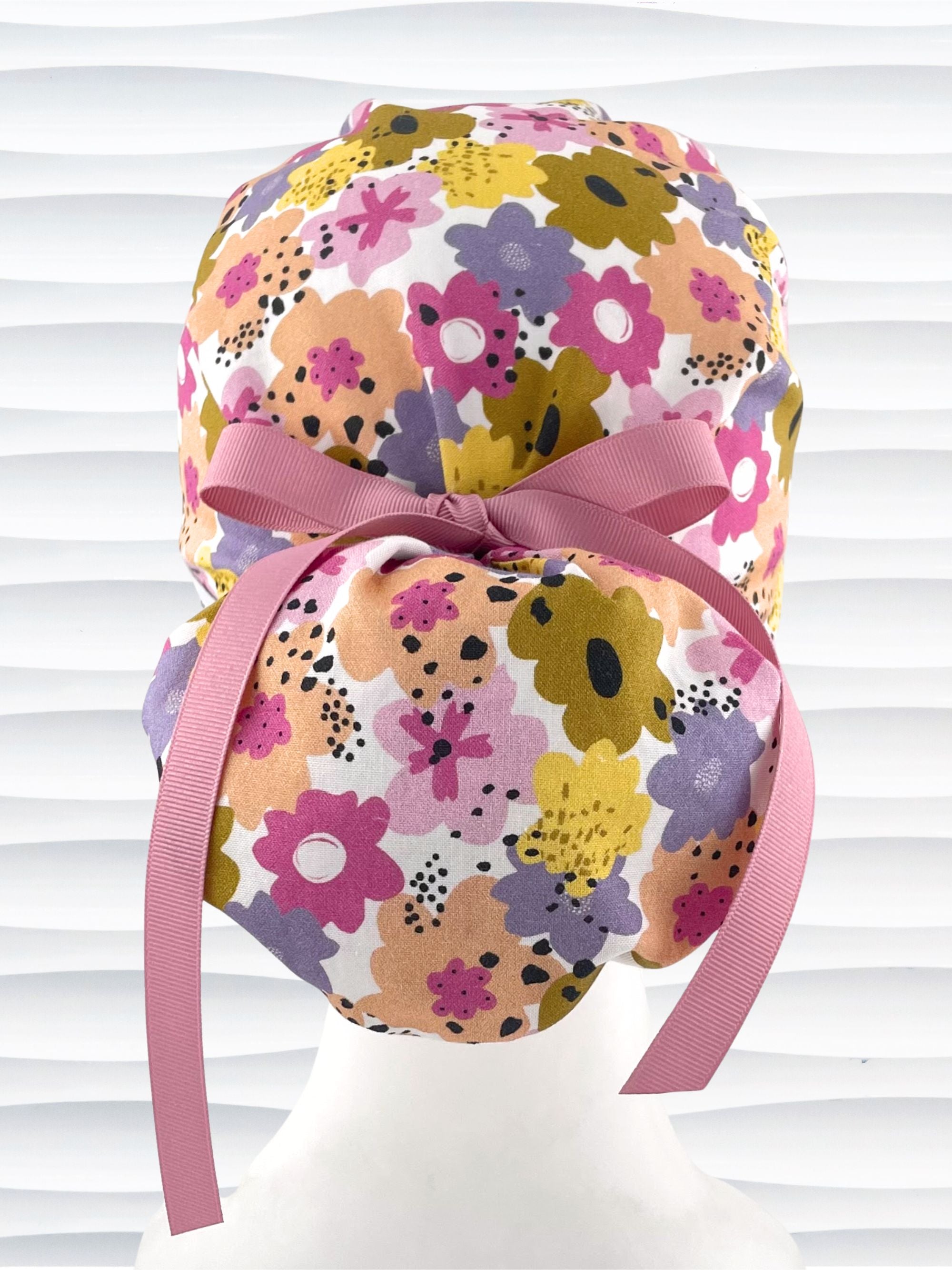 Euro Pony style surgical scrub cap by JensCaps with pink, purple, yellow, and green flowers all over white cotton fabric, tied with a ribbon and displayed on a mannequin head.