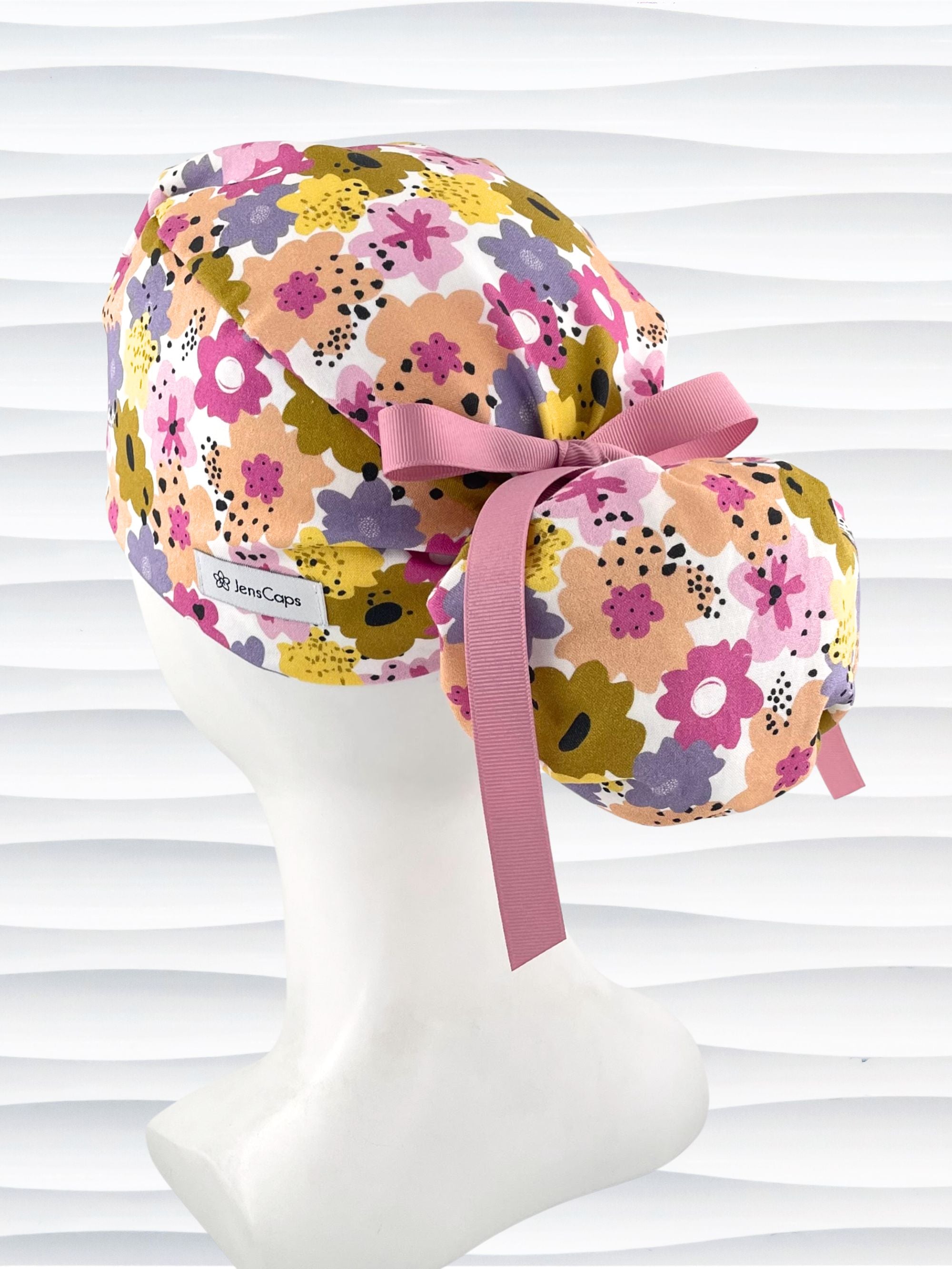 Ponytail style surgical scrub cap by JensCaps with pink, purple, yellow, and green flowers all over white cotton fabric, tied with a ribbon and displayed on a mannequin head.