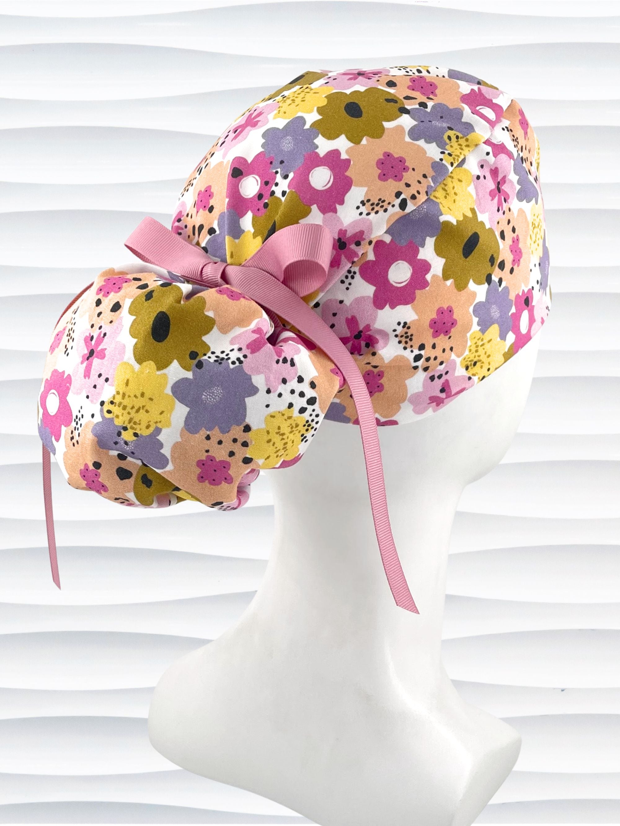 Ponytail style surgical scrub cap by JensCaps with pink, purple, yellow, and green flowers all over white cotton fabric, tied with a ribbon and displayed on a mannequin head.