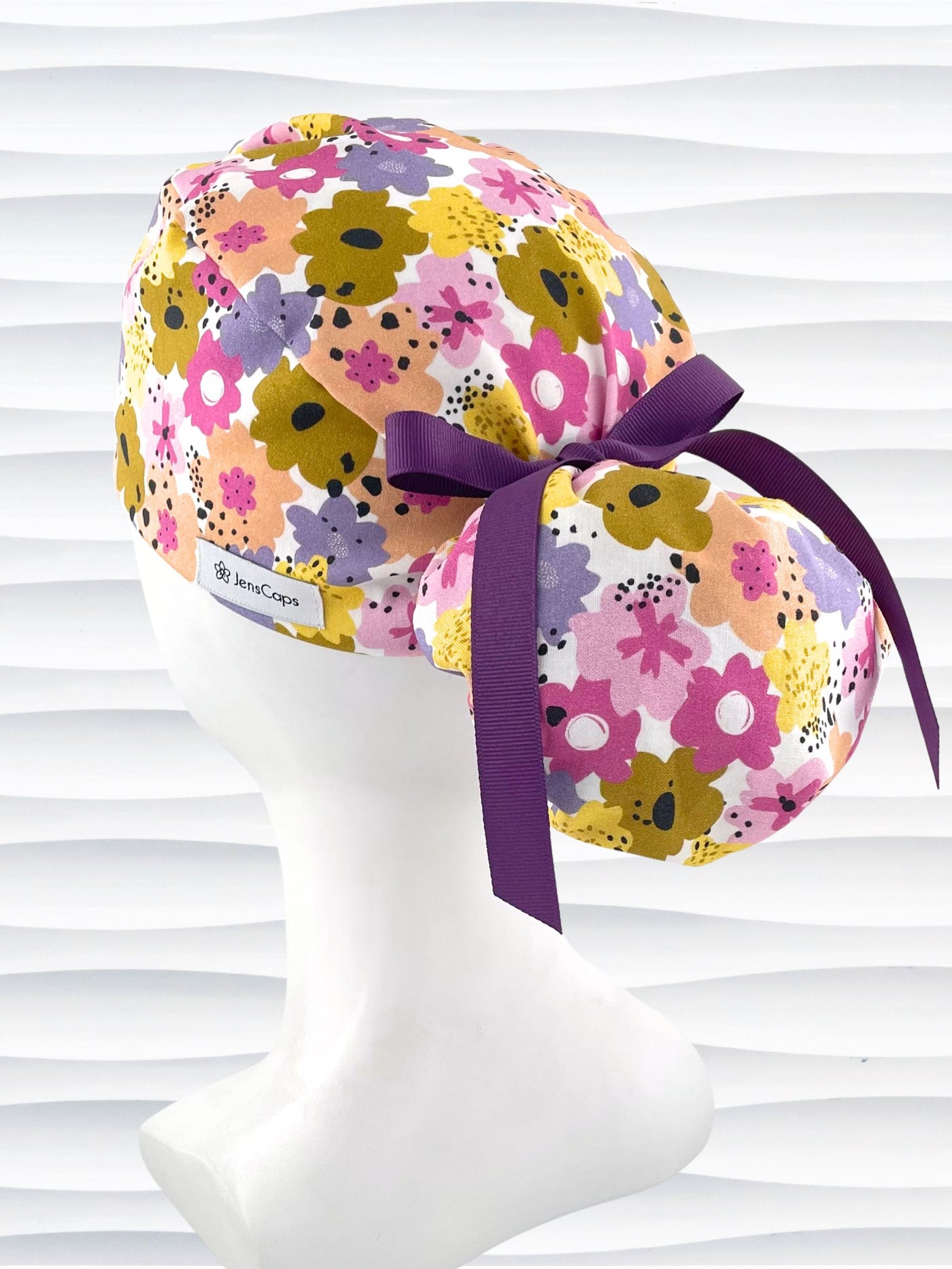 Ponytail style surgical scrub cap by JensCaps with pink, purple, yellow, and green flowers all over white cotton fabric, tied with a ribbon and displayed on a mannequin head.