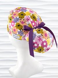 Ponytail style surgical scrub cap by JensCaps with pink, purple, yellow, and green flowers all over white cotton fabric, tied with a ribbon and displayed on a mannequin head.