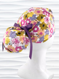 Ponytail style surgical scrub cap by JensCaps with pink, purple, yellow, and green flowers all over white cotton fabric, tied with a ribbon and displayed on a mannequin head.