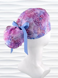 Ponytail style surgical scrub cap by JensCaps with small daisies in pink, blue, and purple all over cotton fabric, tied with a ribbon, and displayed on a mannequin head
