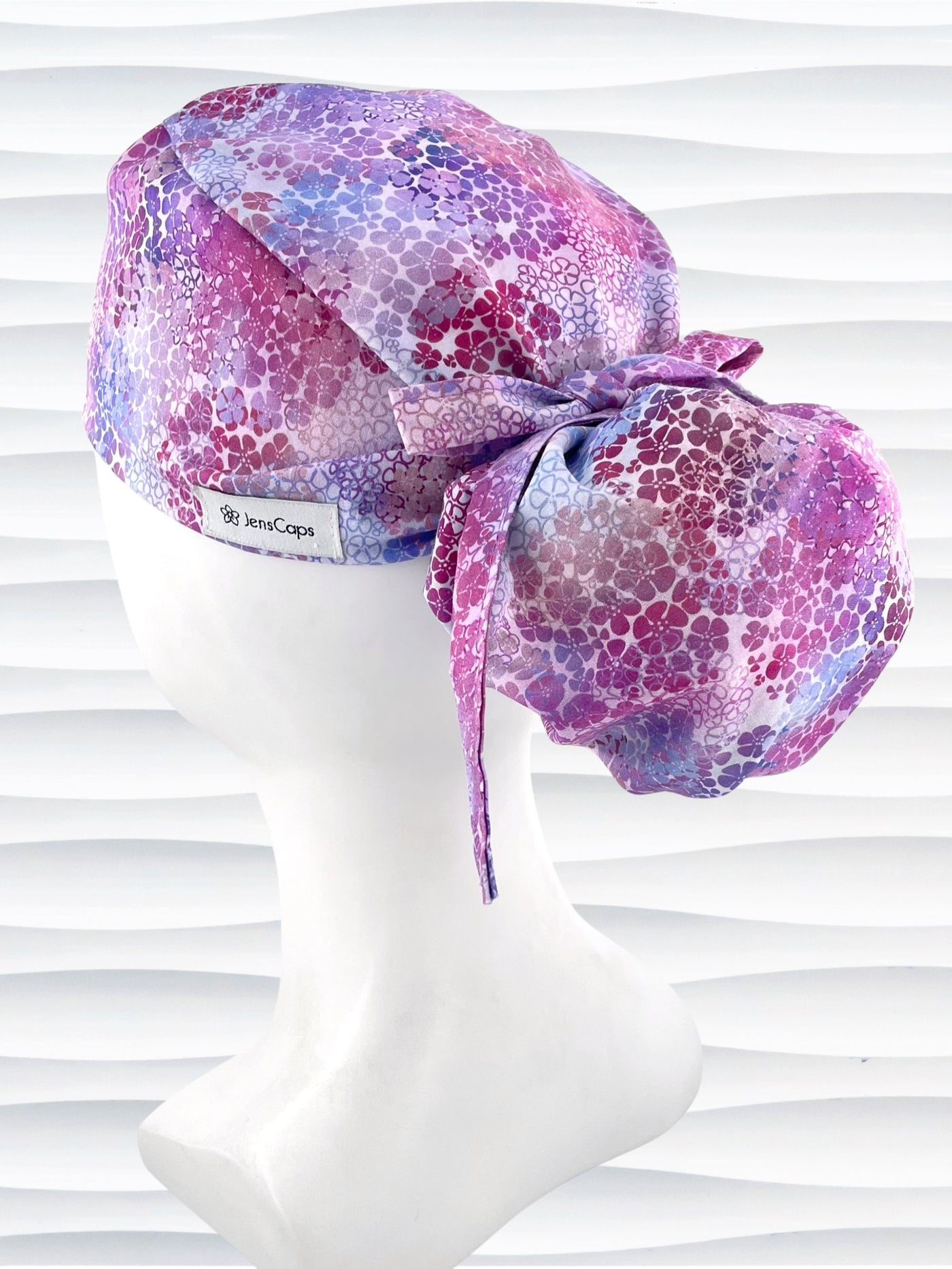 Ponytail style surgical scrub cap by JensCaps with small daisies in pink, blue, and purple all over cotton fabric, tied with a ribbon, and displayed on a mannequin head