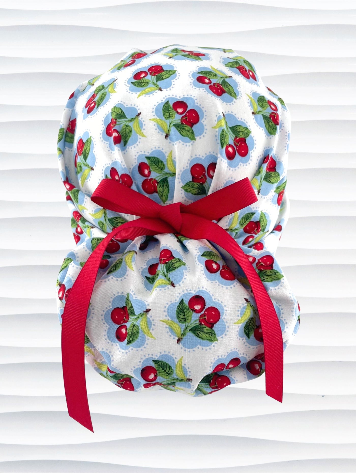Euro Pony style surgical scrub cap by JensCaps with red cherries blue polka dots on white cotton fabric, finished with a ribbon tie, and displayed on a mannequin head.