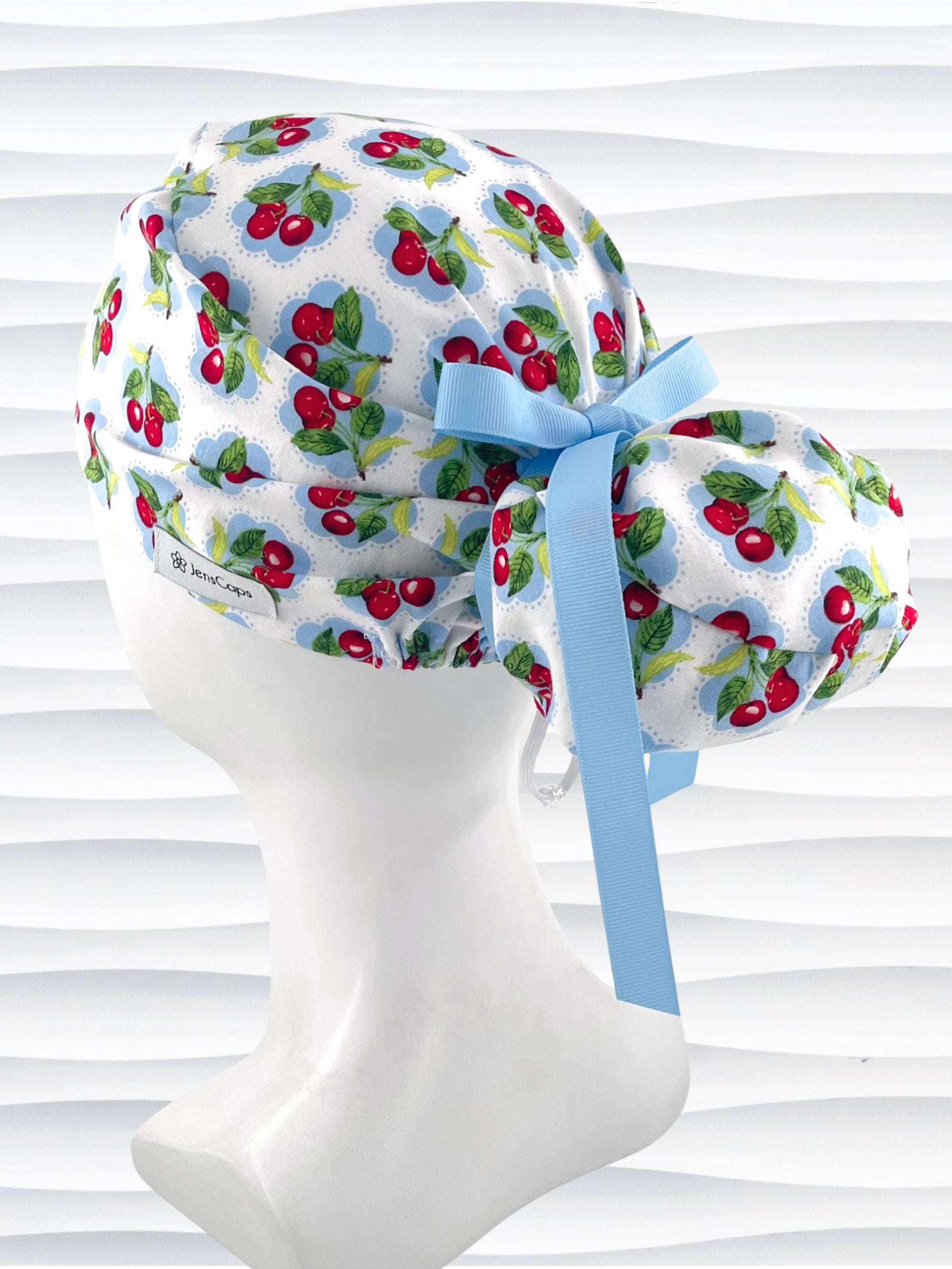 Euro Pony style surgical scrub cap by JensCaps with red cherries blue polka dots on white cotton fabric, finished with a ribbon tie, and displayed on a mannequin head.