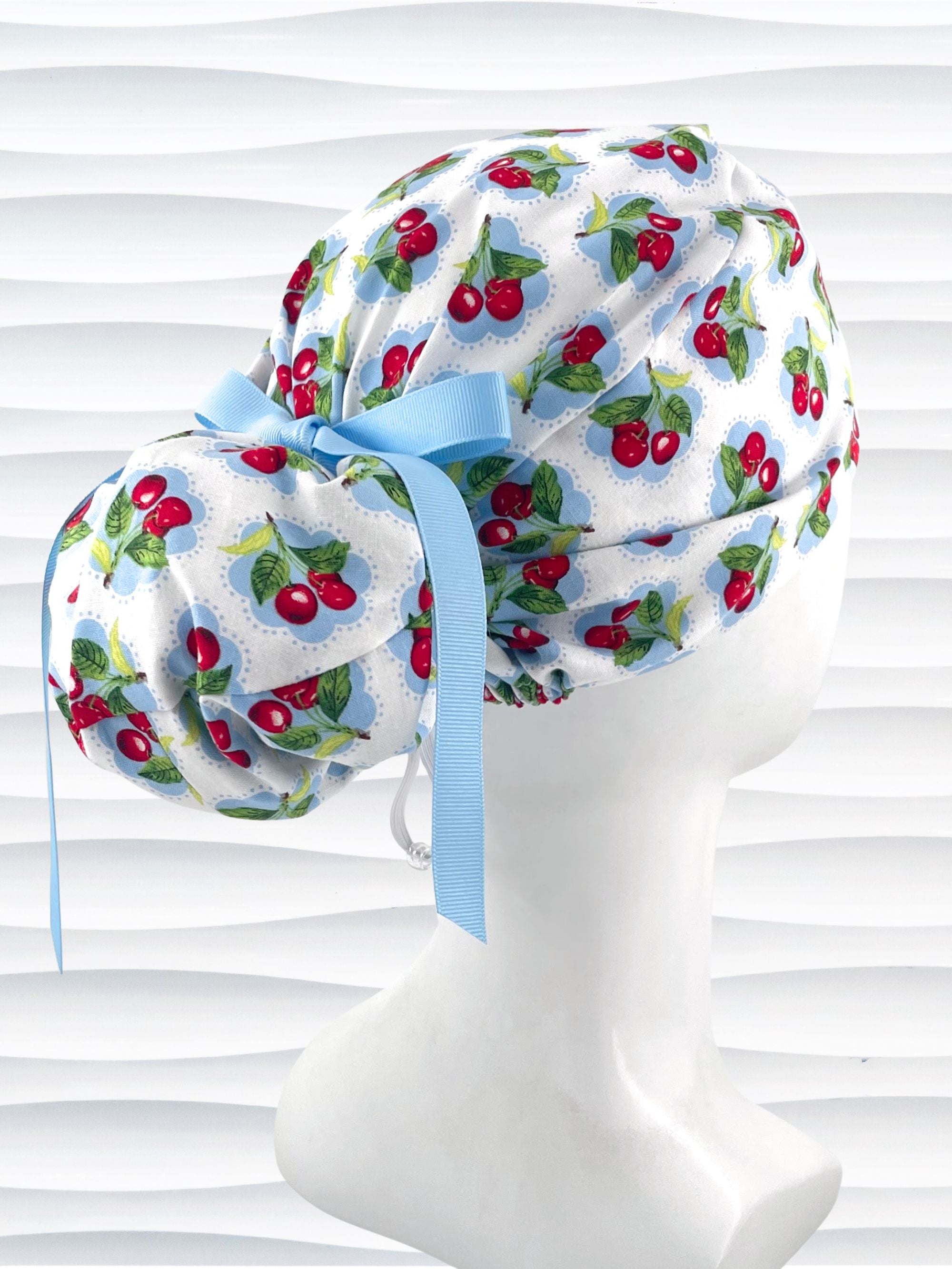 Euro Pony style surgical scrub cap by JensCaps with red cherries blue polka dots on white cotton fabric, finished with a ribbon tie, and displayed on a mannequin head.