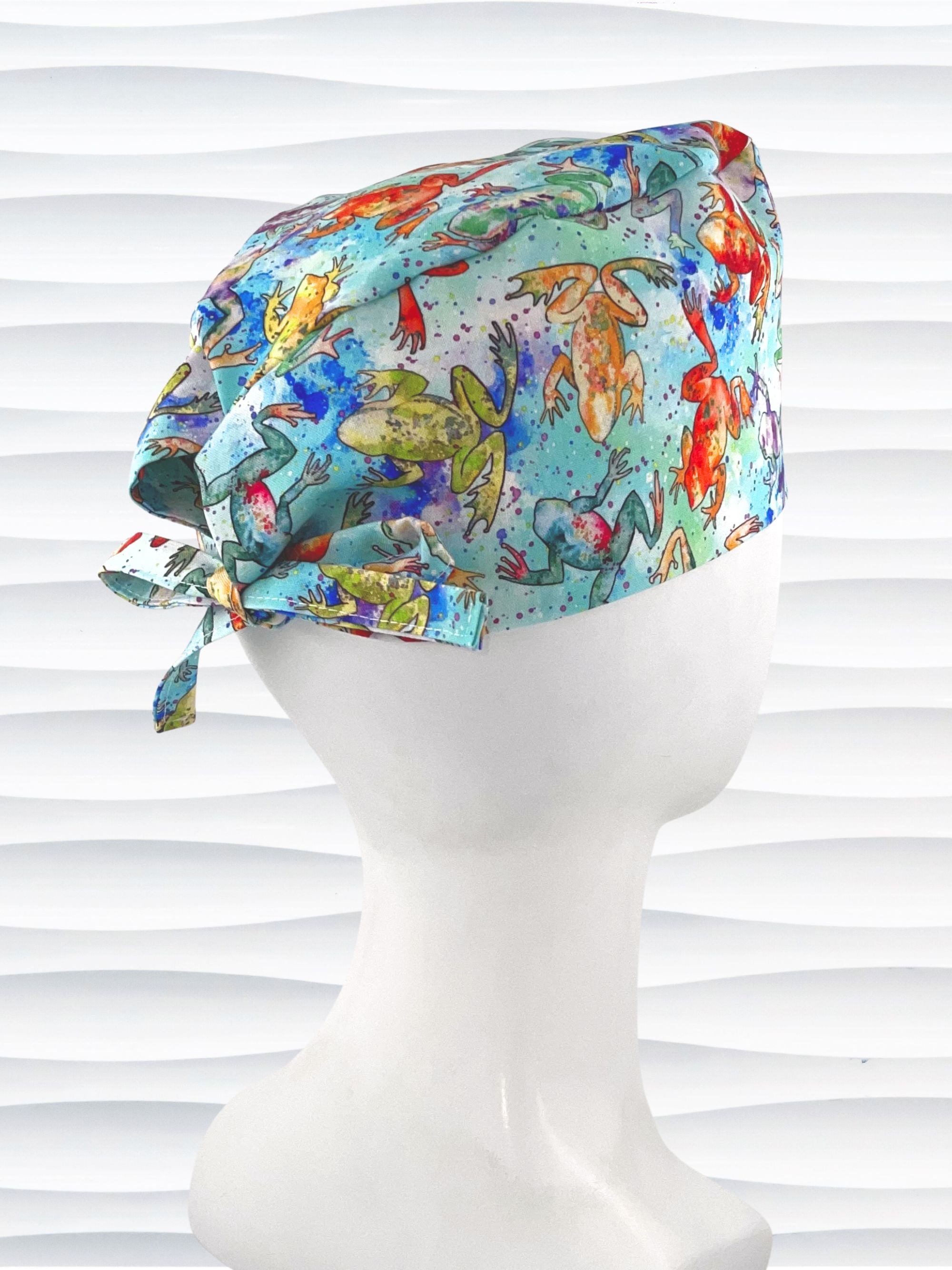 Surgical scrub cap by JensCaps in an adjustable tie-back style with a colorful watercolor frog print in orange, purple, green, and blue on an aqua background and displayed on a mannequin head 