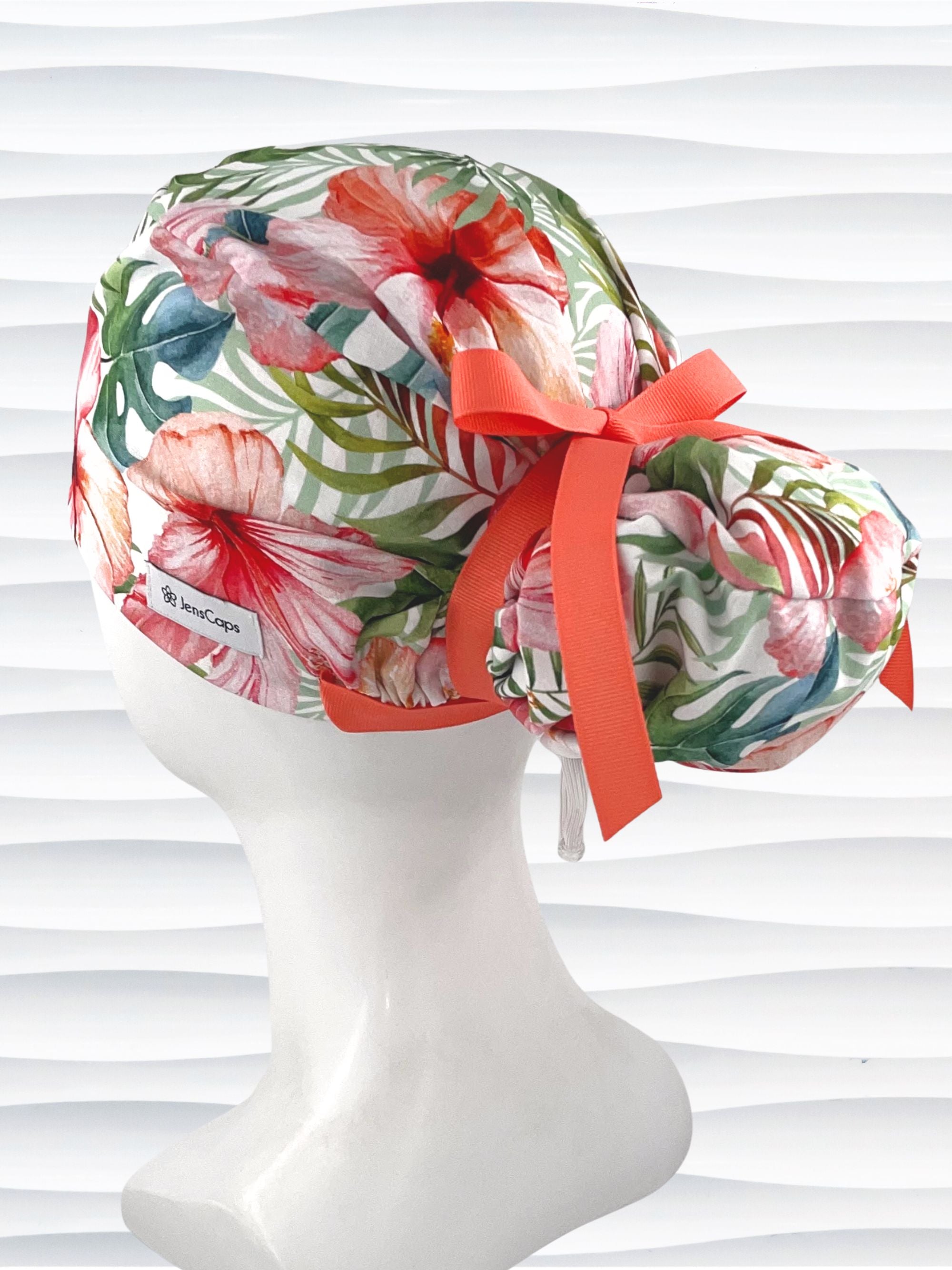 Euro Pony style surgical scrub cap by JensCaps with tropical hibiscus flowers and palm fronds all over white cotton fabric, finished with a grosgrain ribbon tie, and displayed on a mannequin head