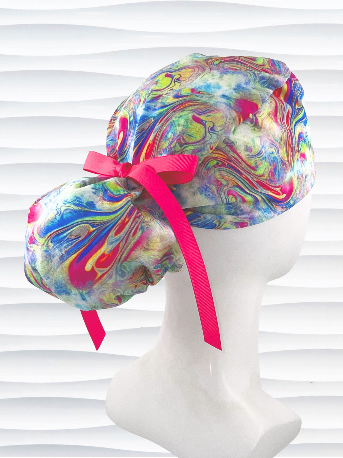 Ponytail style surgical scrub cap by JensCaps with an abstract marbled swirl print in blue, pink, yellow, and green, finished with a grosgrain ribbon, and displayed on a white mannequin head.