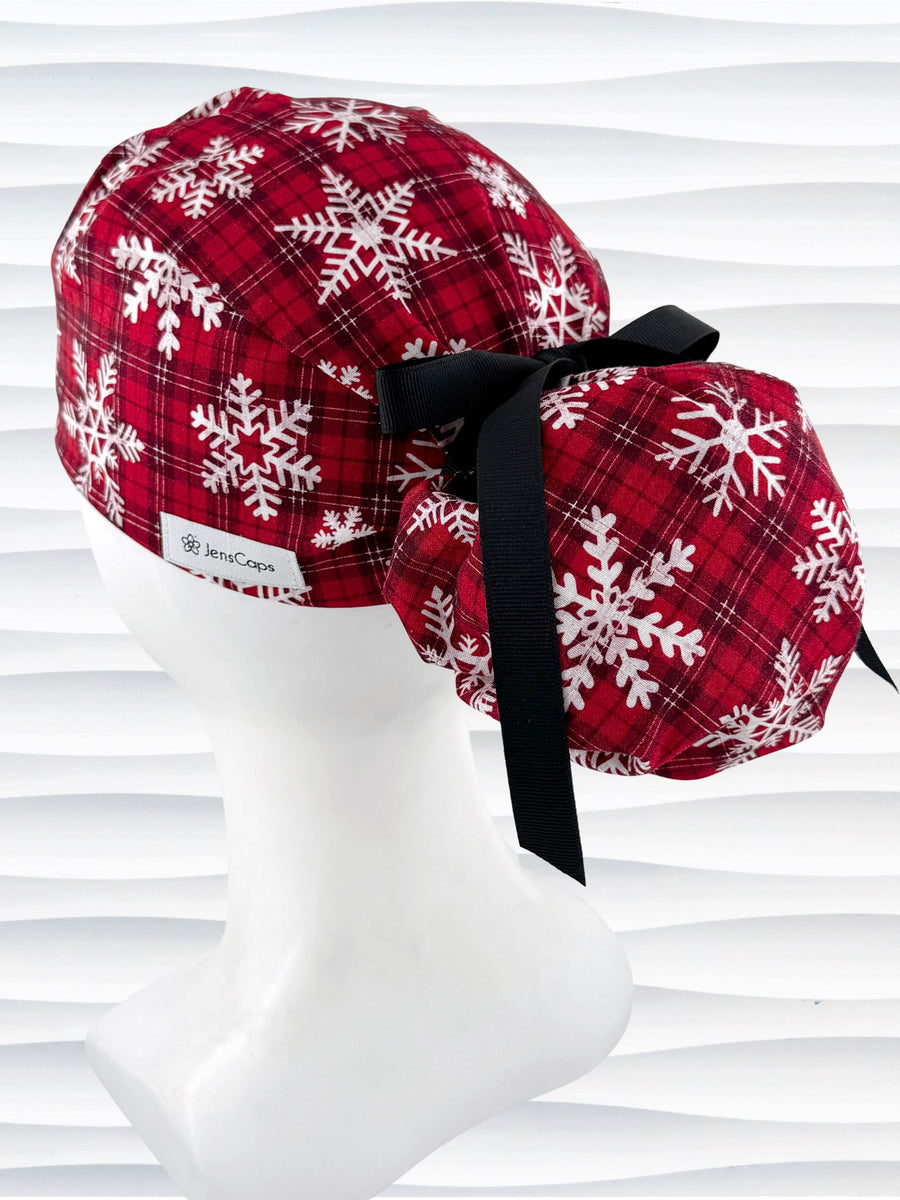 Red plaid ponytail scrub cap with white snowflake patterns and a black ribbon tie at the back, displayed on a white mannequin head against a wavy white background.