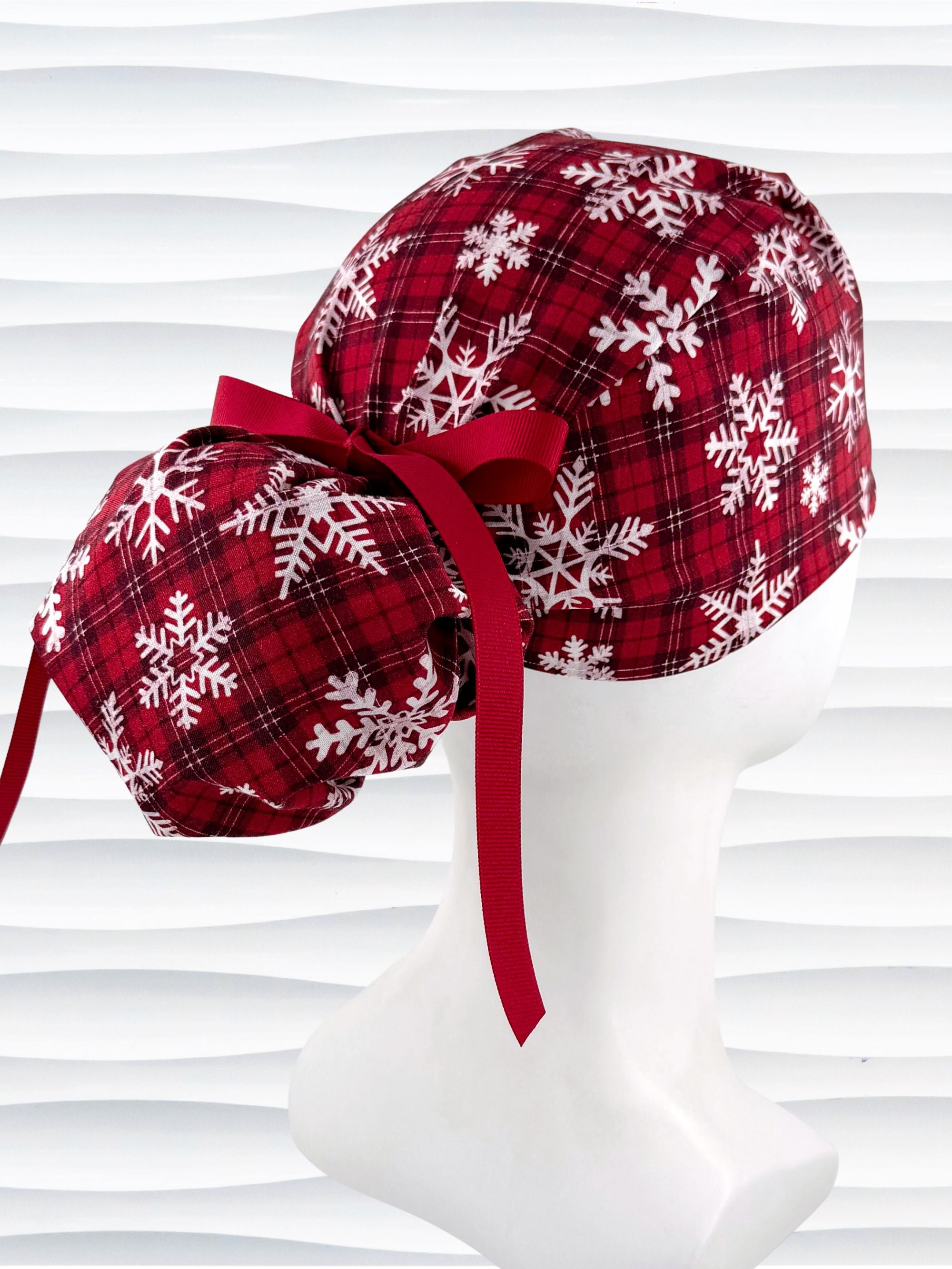 Red plaid ponytail scrub cap with white snowflake patterns and a red ribbon tie at the back, displayed on a white mannequin head against a wavy white background.