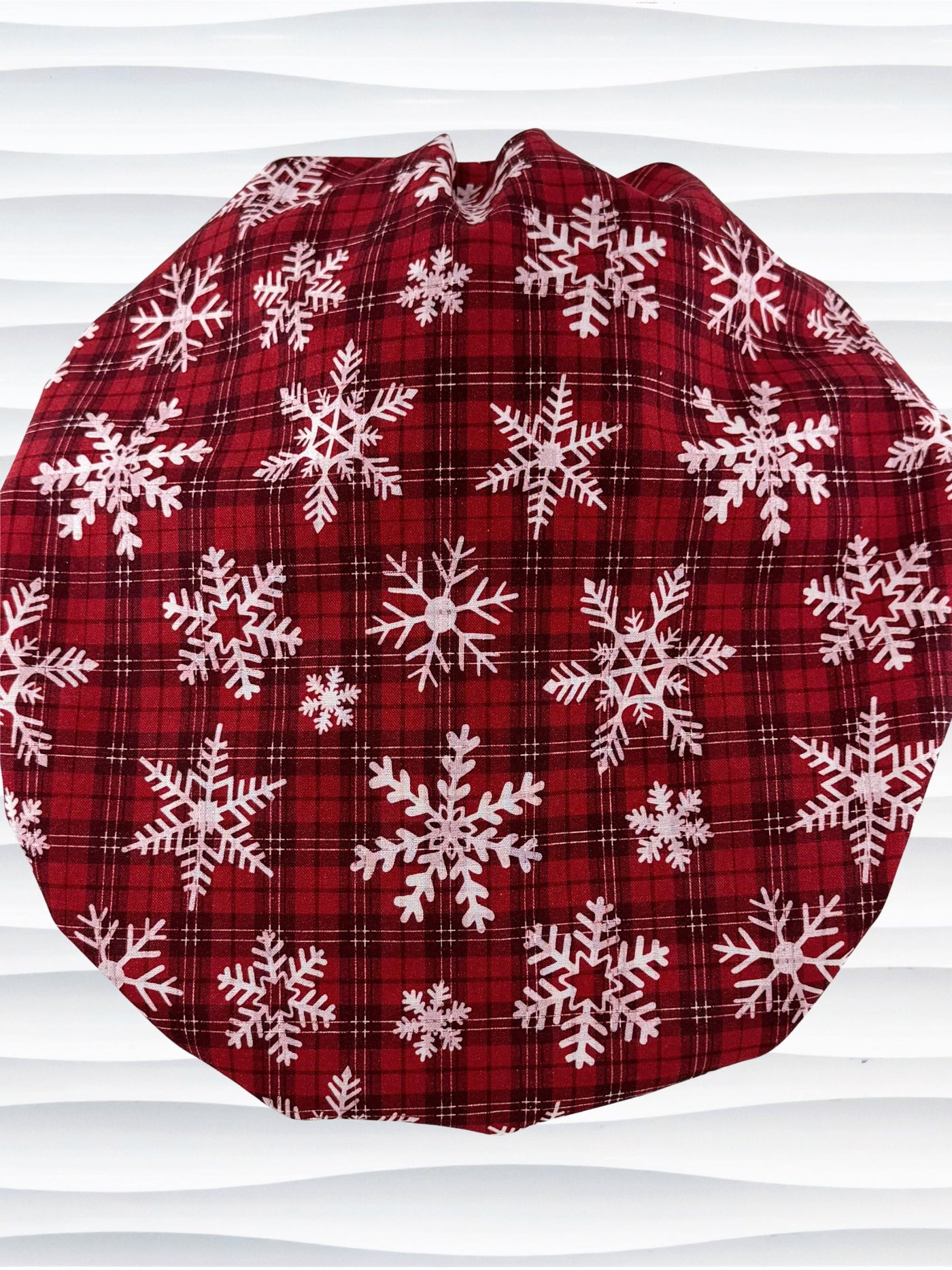 Red plaid bouffant scrub cap by jenscaps with white snowflake patterns displayed on a white mannequin head against a wavy white background.