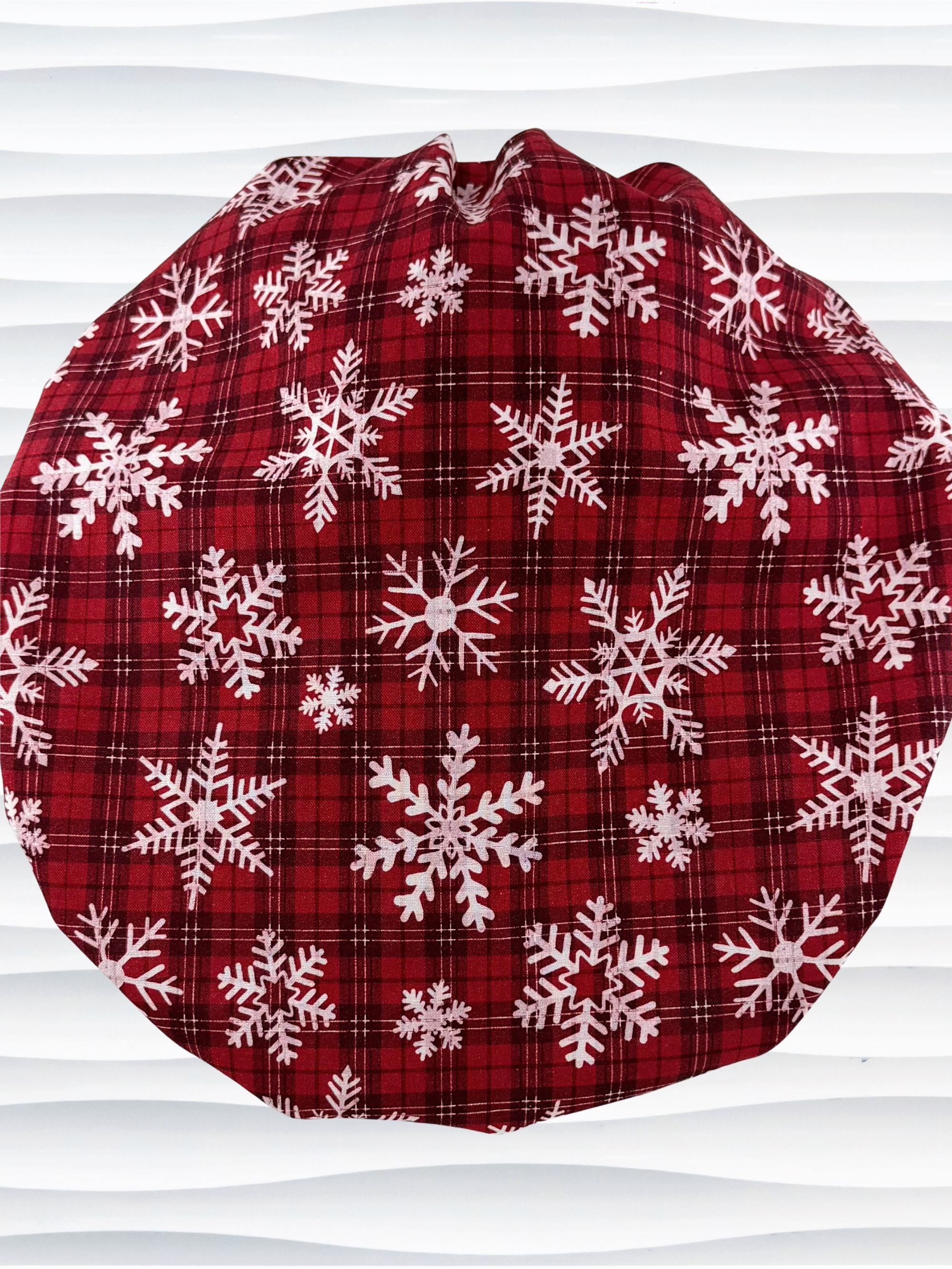 Red plaid bouffant scrub cap by jenscaps with white snowflake patterns displayed on a white mannequin head against a wavy white background.