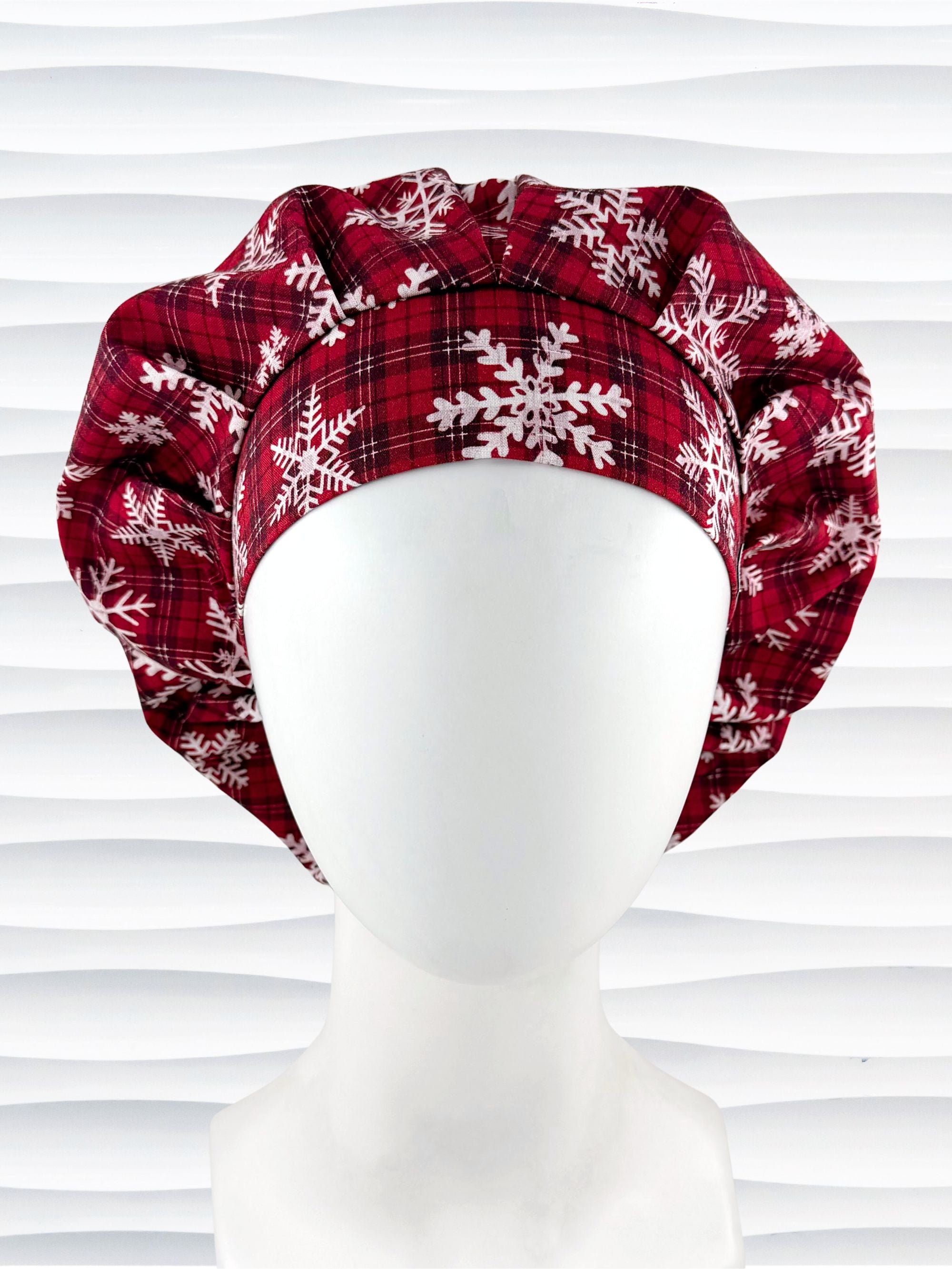Red plaid bouffant scrub cap by jenscaps with white snowflake patterns displayed on a white mannequin head against a wavy white background.