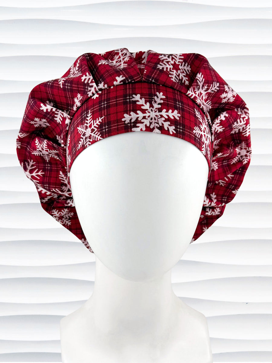 Red plaid bouffant scrub cap by jenscaps with white snowflake patterns displayed on a white mannequin head against a wavy white background.