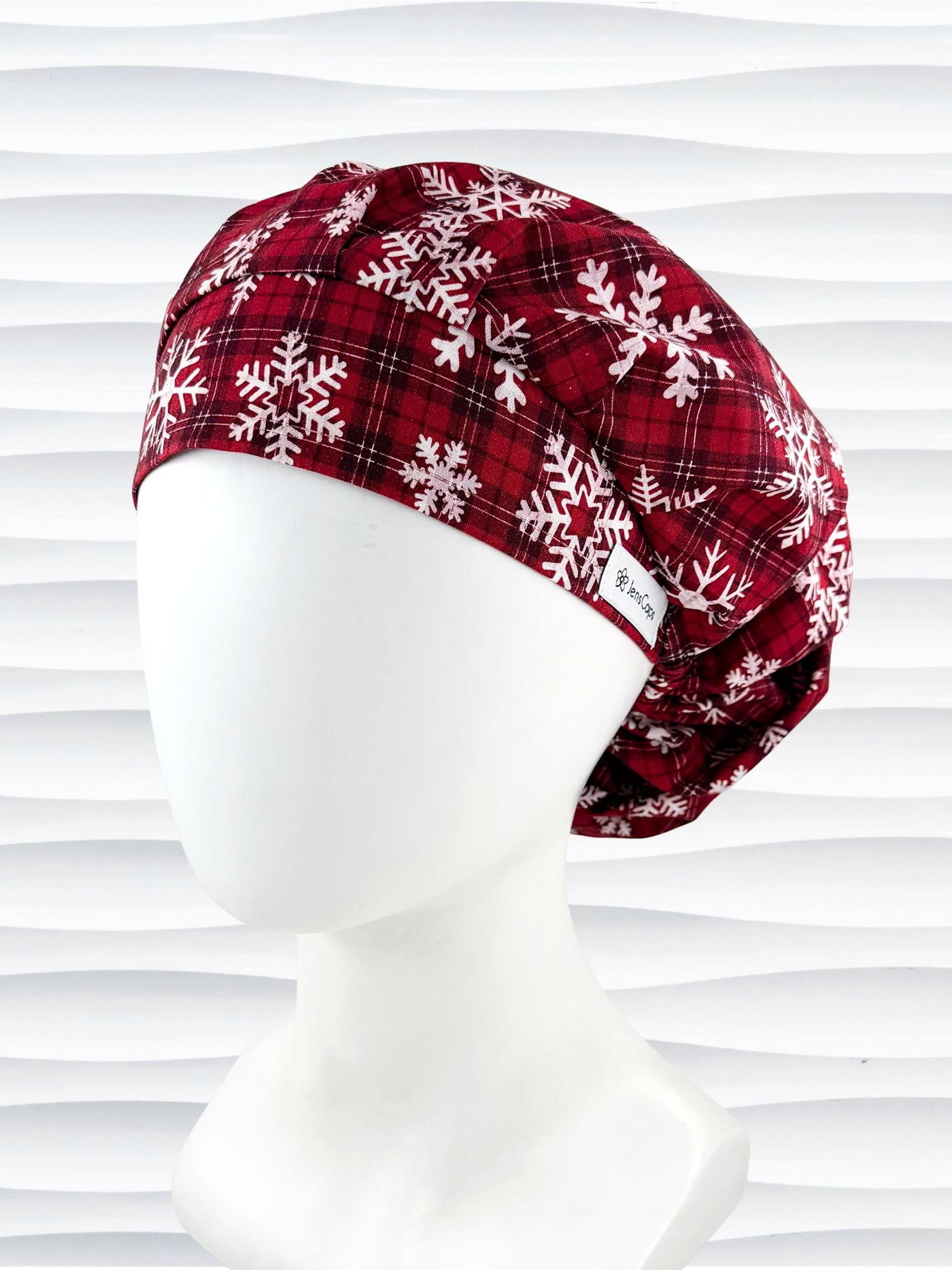 Red plaid bouffant scrub cap by jenscaps with white snowflake patterns displayed on a white mannequin head against a wavy white background.
