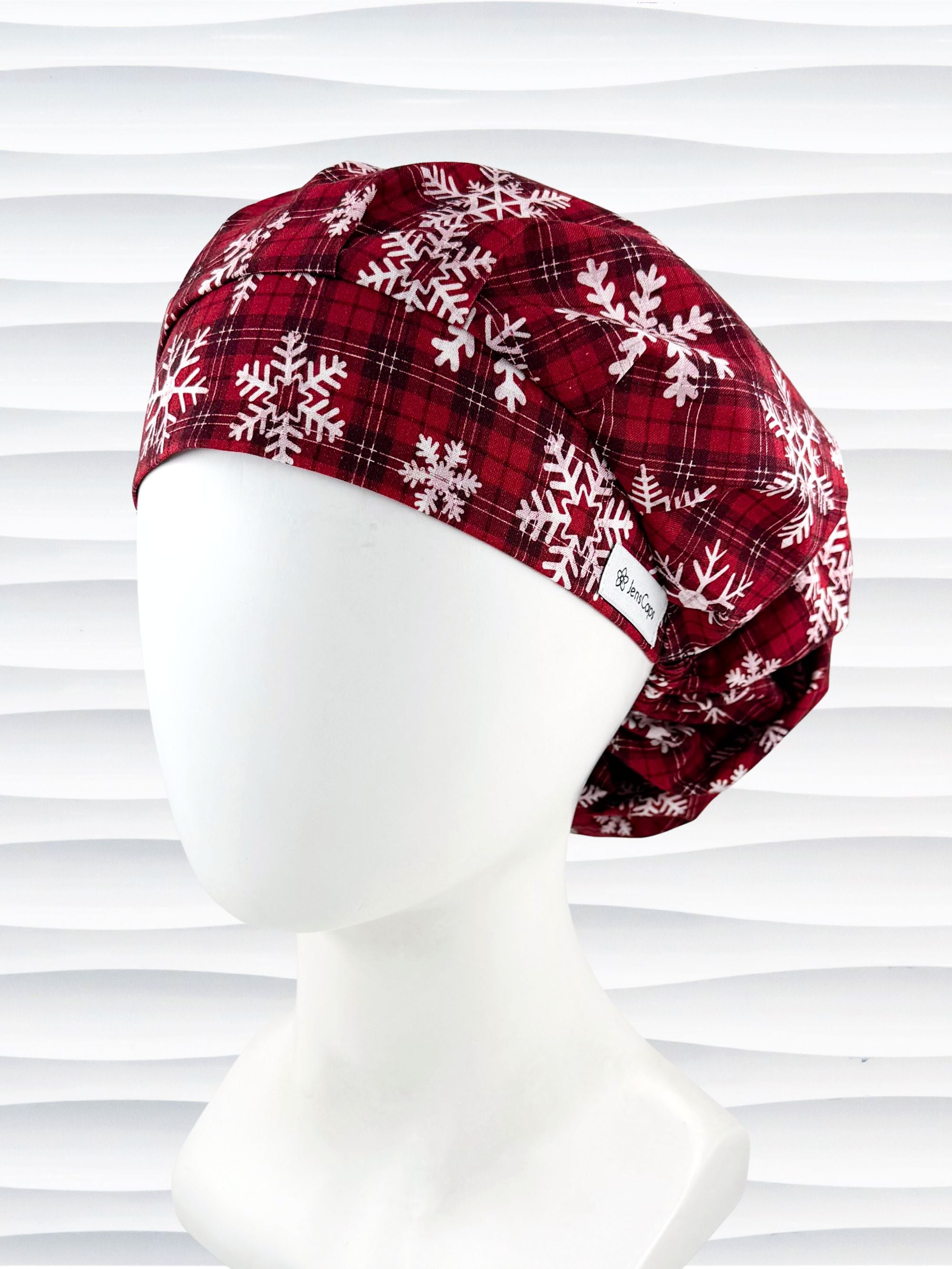Red plaid bouffant scrub cap by jenscaps with white snowflake patterns displayed on a white mannequin head against a wavy white background.