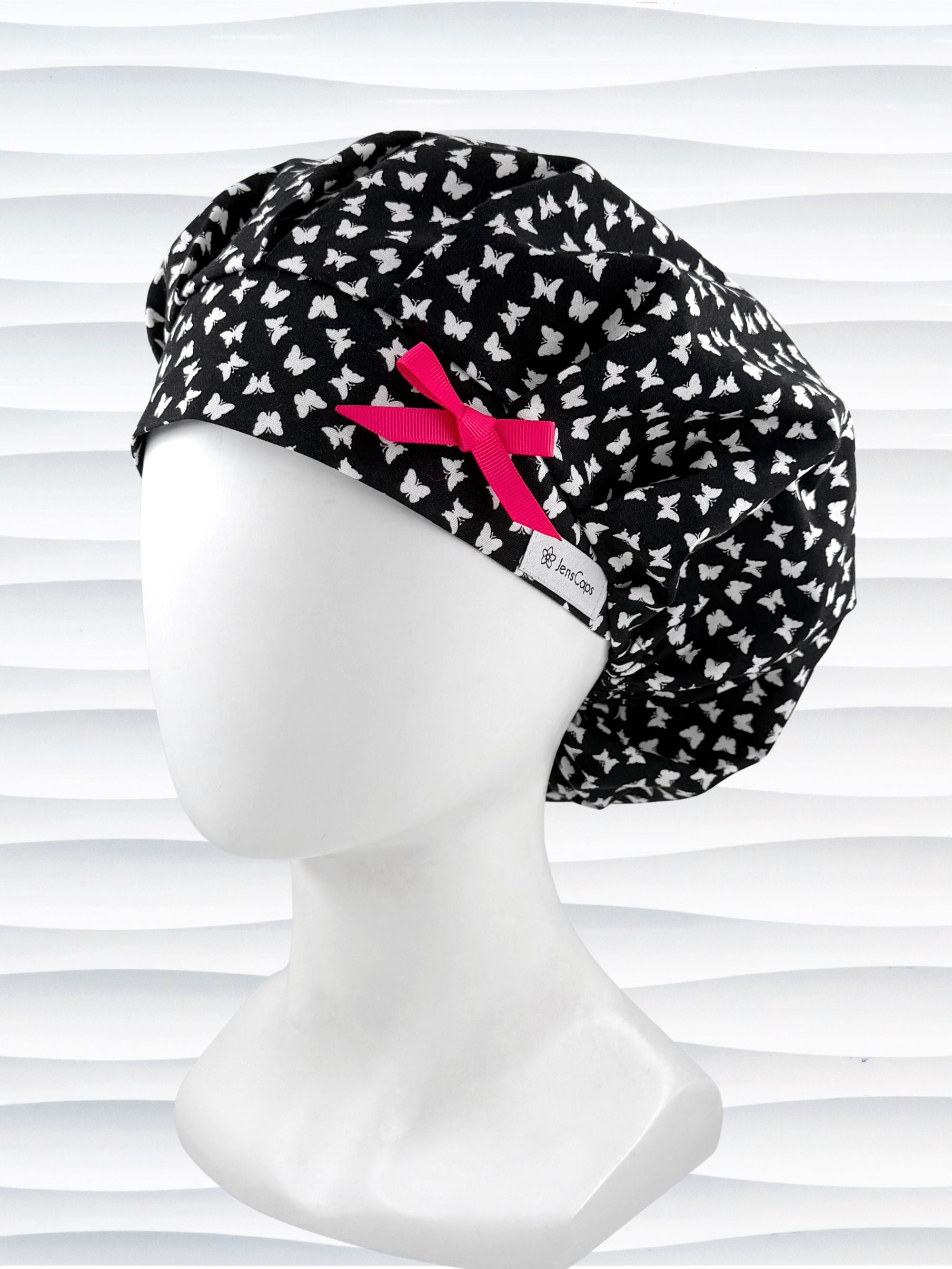 Bouffant surgical scrub cap by JensCaps with white butterflies all over black cotton fabric and shown with a hot pink bow on the front