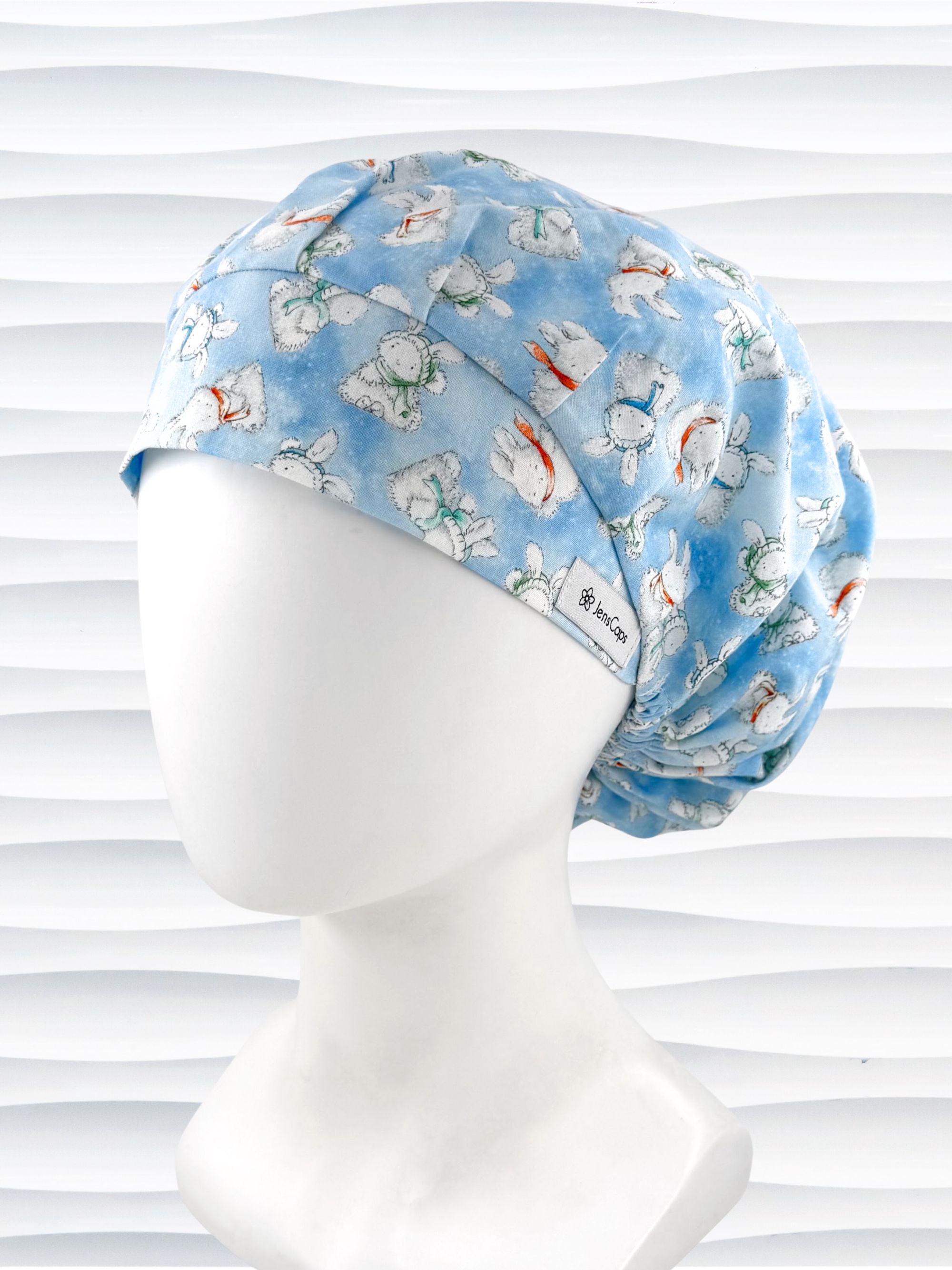 Bouffant style surgical scrub cap by JensCaps with white bunnies wearing beanies and scarves tossed all over this light blue cotton fabric and displayed on a white mannequin head