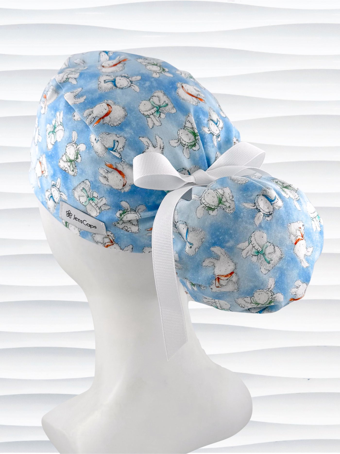 Ponytail style surgical scrub cap by JensCaps with white bunnies wearing beanies and scarves tossed all over this light blue cotton fabric, finished with a ribbon tie, and displayed on a white mannequin head