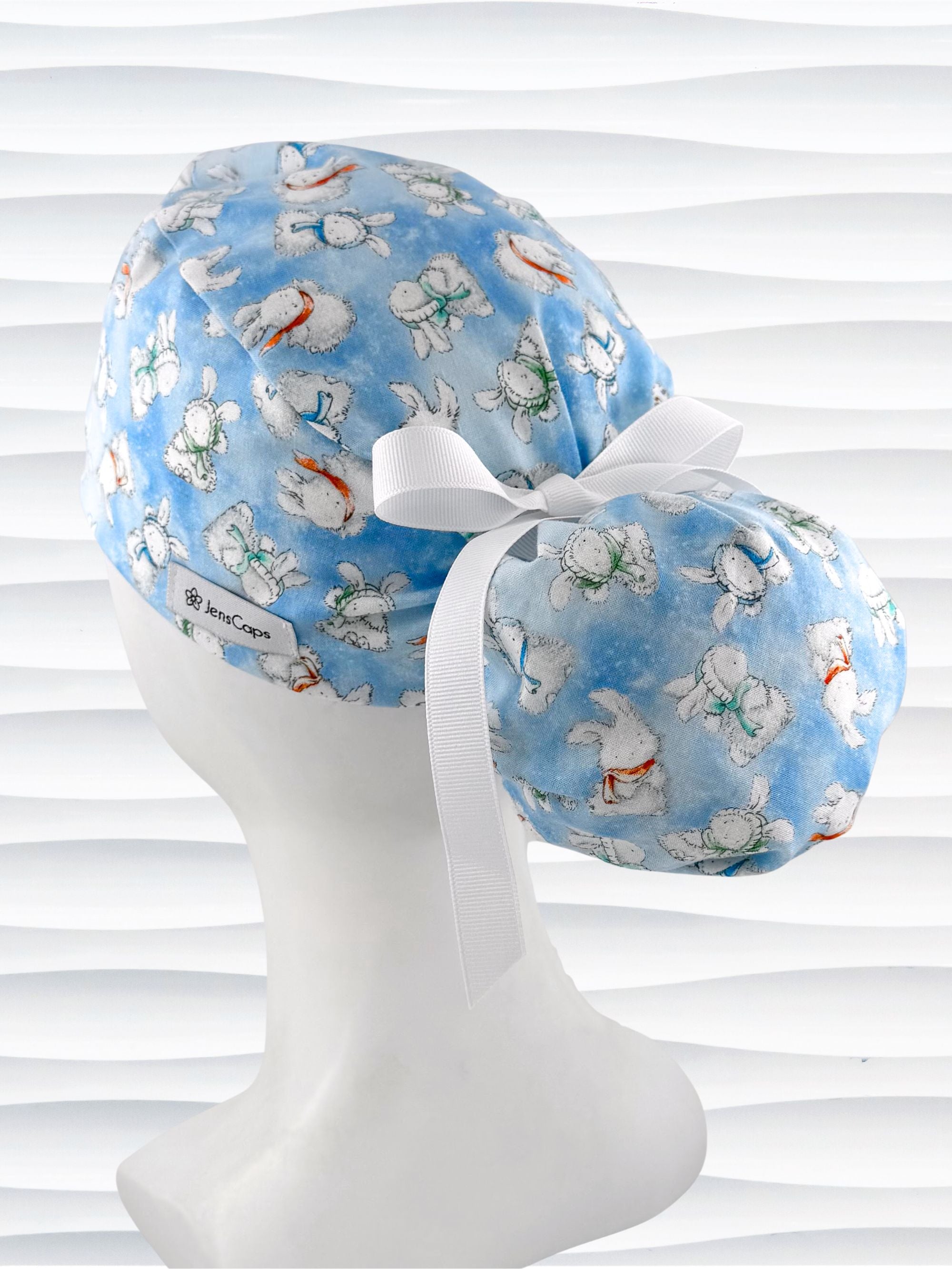 Ponytail style surgical scrub cap by JensCaps with white bunnies wearing beanies and scarves tossed all over this light blue cotton fabric, finished with a ribbon tie, and displayed on a white mannequin head