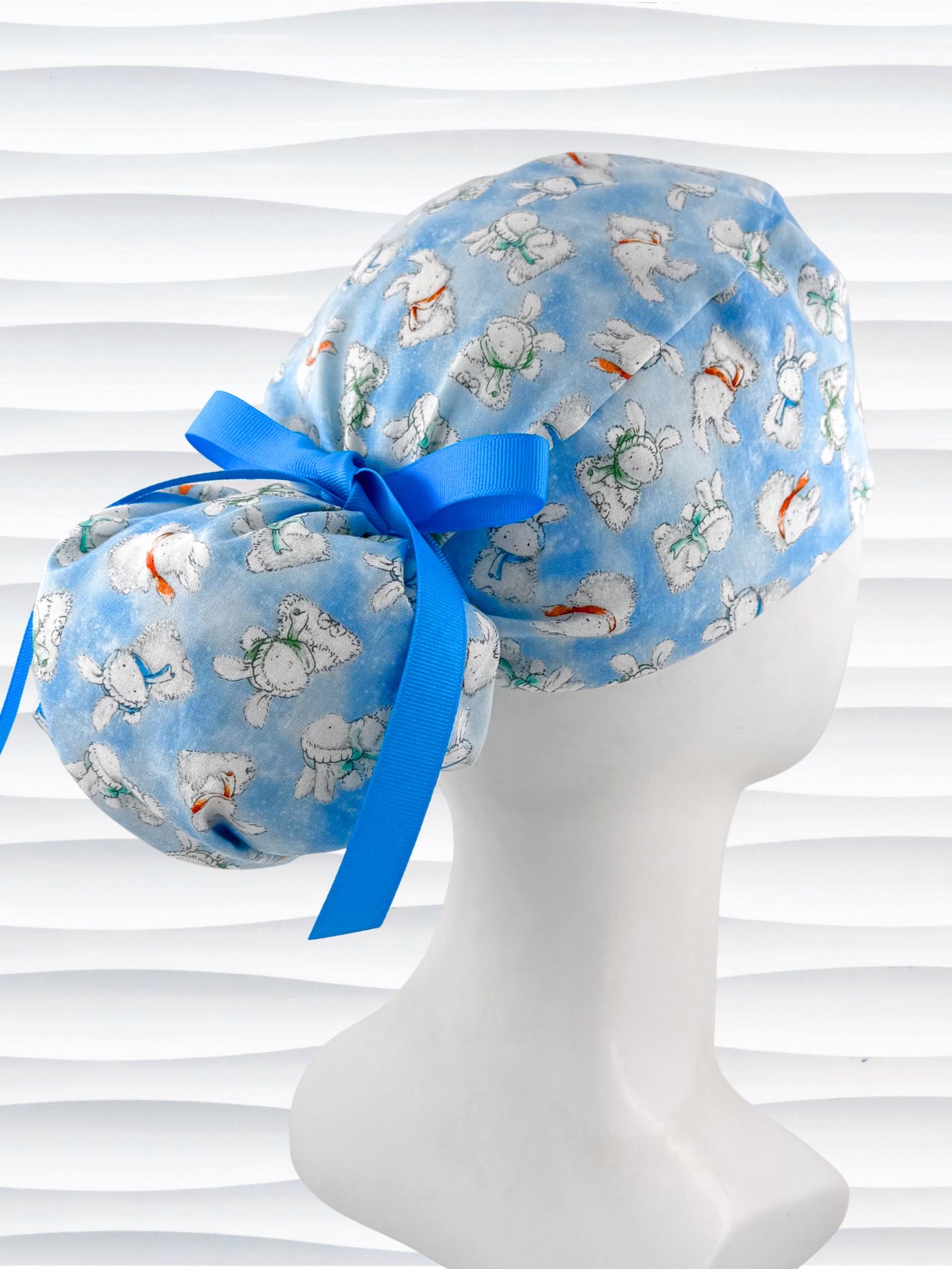 Ponytail style surgical scrub cap by JensCaps with white bunnies wearing beanies and scarves tossed all over this light blue cotton fabric, finished with a ribbon tie, and displayed on a white mannequin head