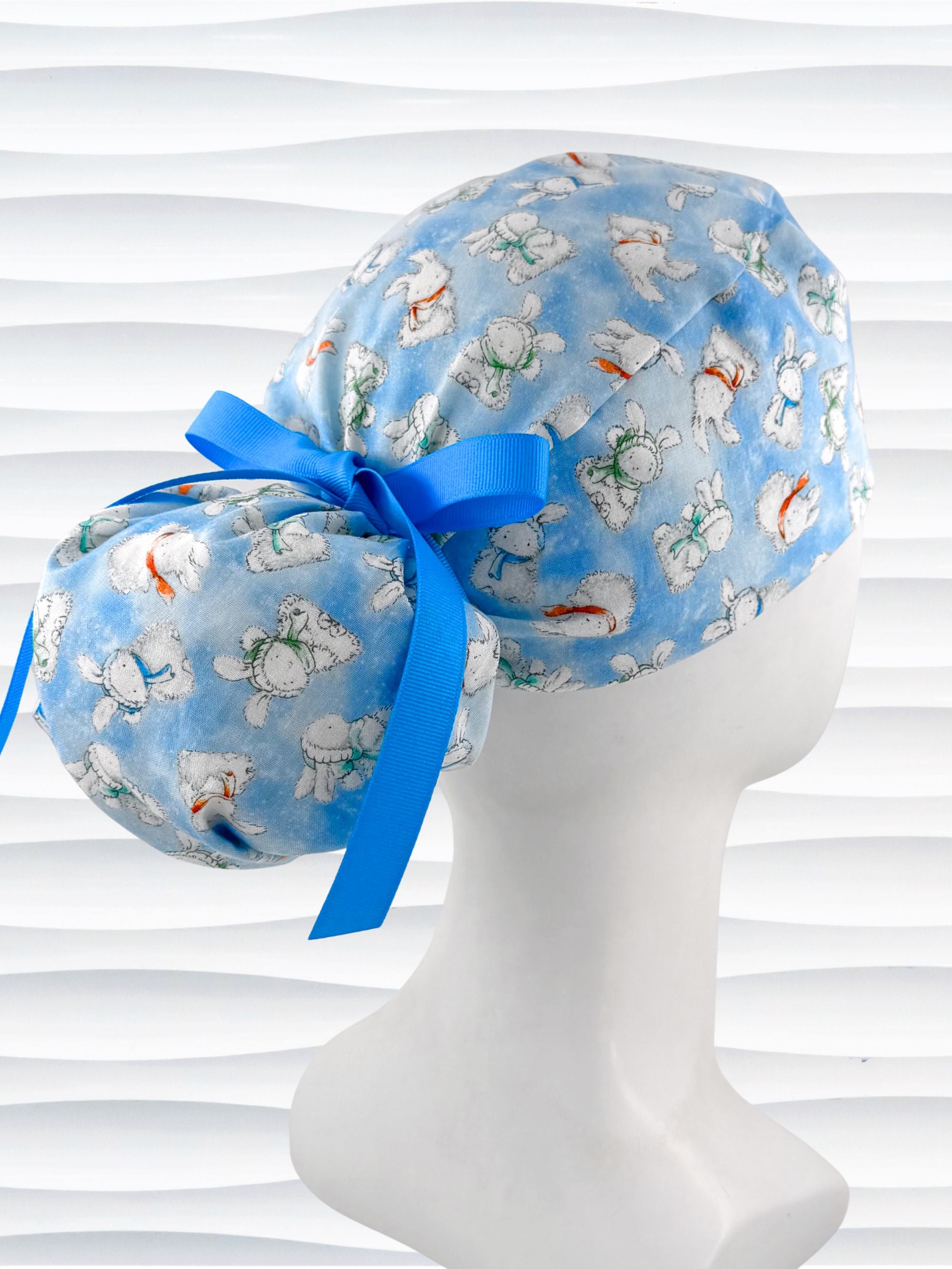 Ponytail style surgical scrub cap by JensCaps with white bunnies wearing beanies and scarves tossed all over this light blue cotton fabric, finished with a ribbon tie, and displayed on a white mannequin head